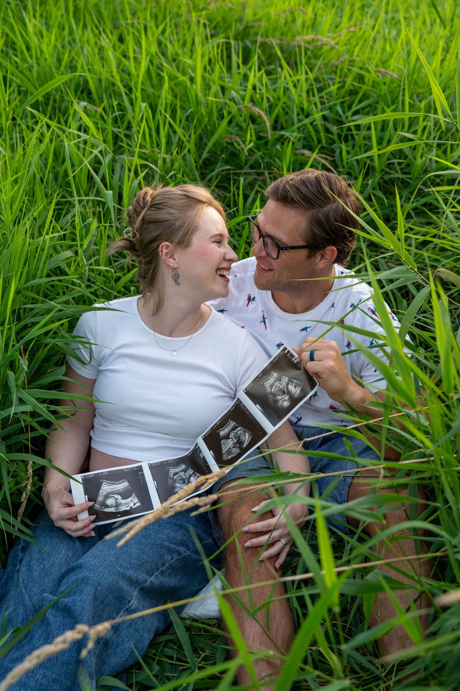Happy couple sitting in lush green grass holding ultrasound photos, smiling at each other during pregnancy announcement.