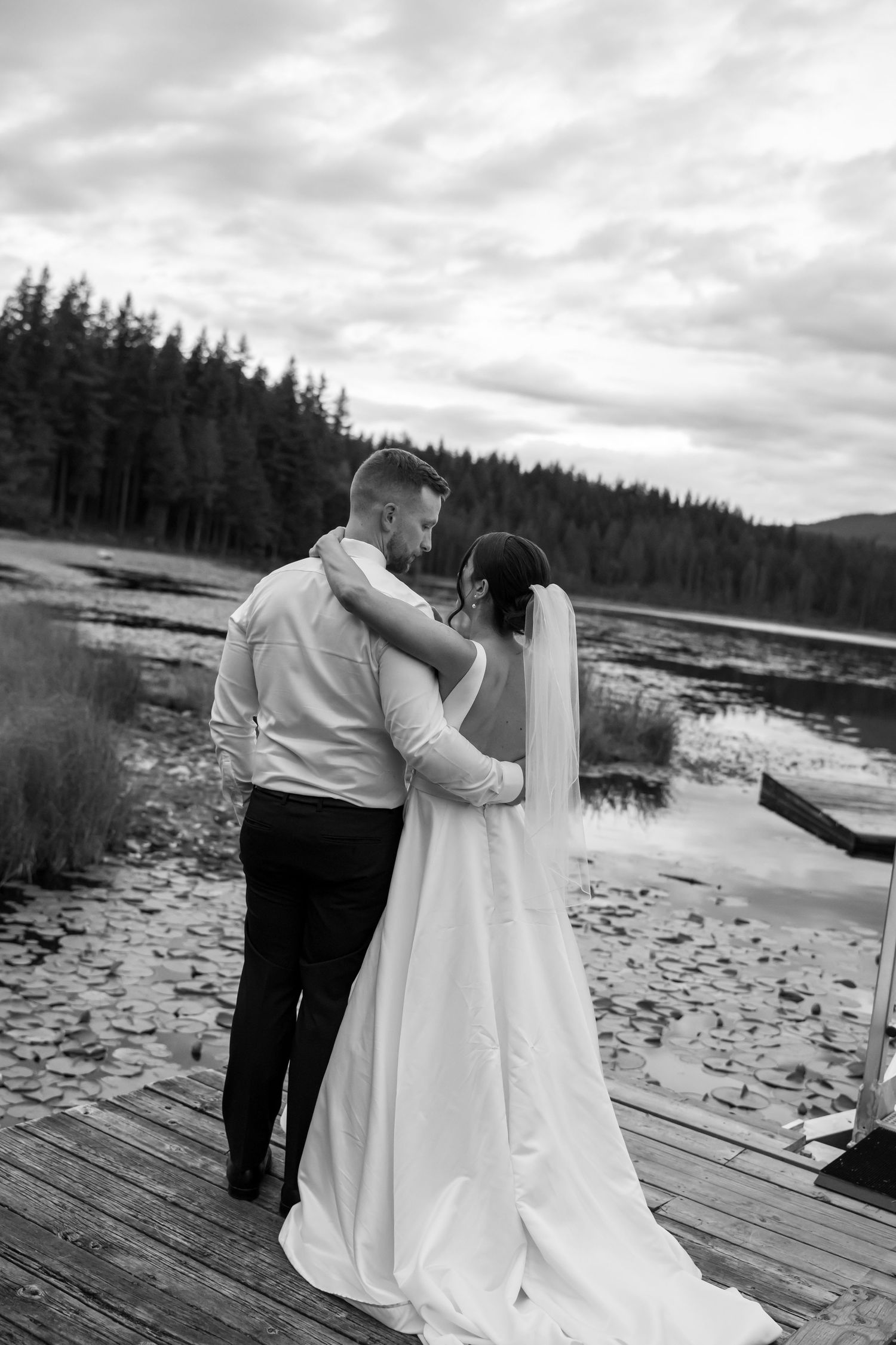 Black and white photo of a bride and groom embracing on a wooden dock by a lake surrounded by pine trees.