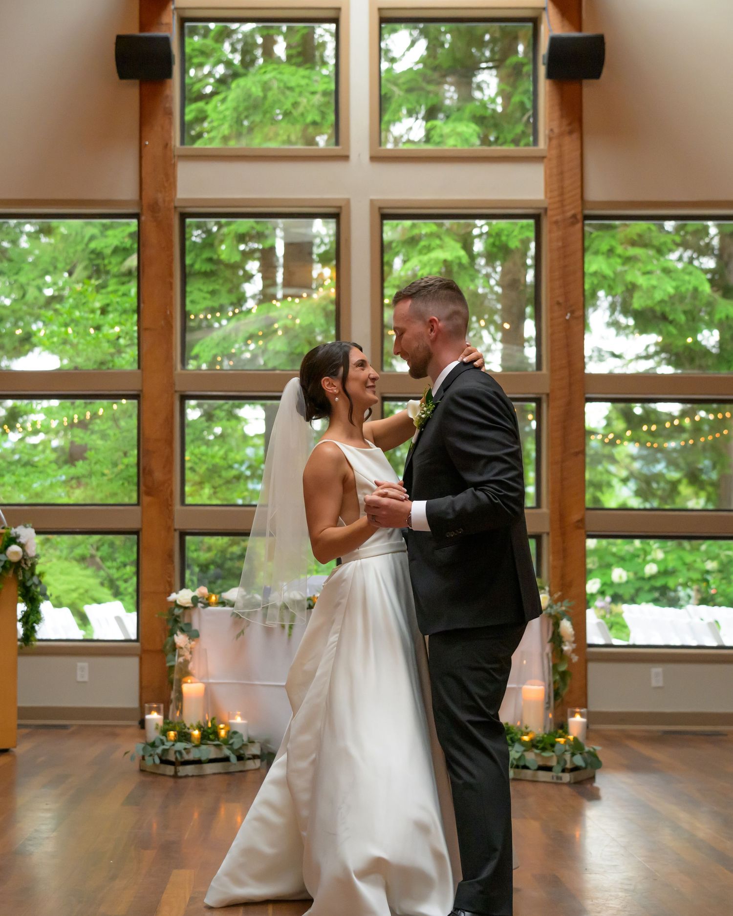 Bride and groom share first dance in elegant wood-framed venue with large windows and lush greenery backdrop.