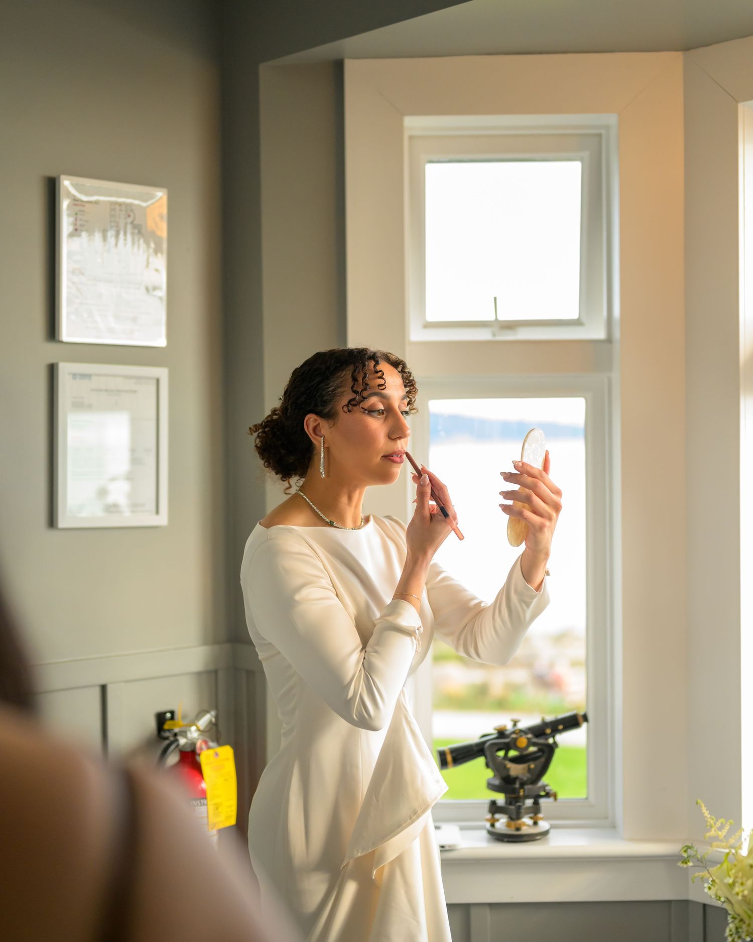 Person in white robe applying makeup near bright window in modern bathroom.