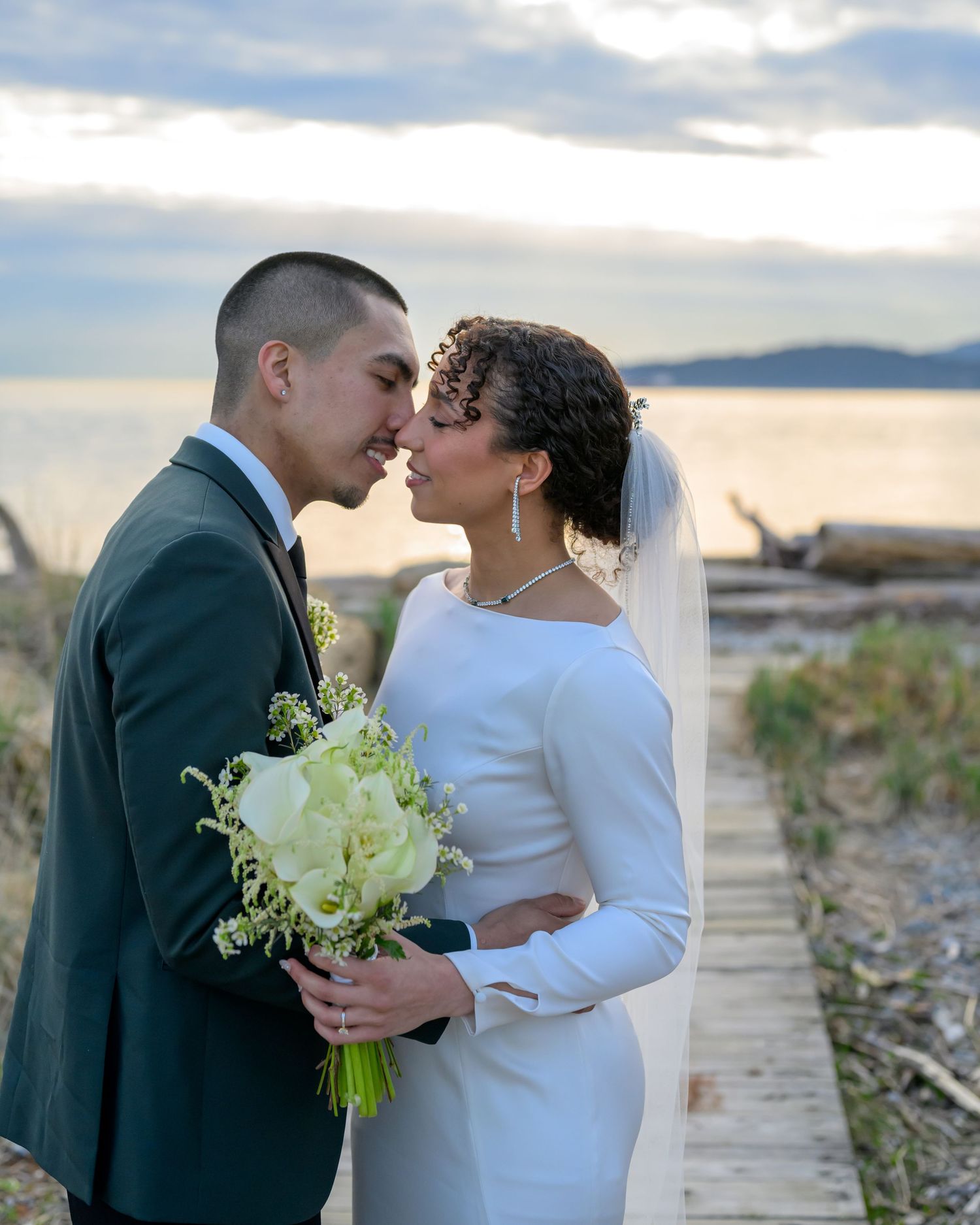Intimate wedding portrait with couple holding white bouquet against sunset coastal backdrop.