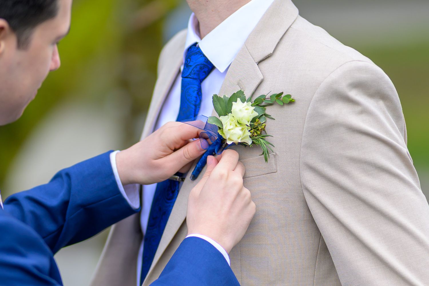 Groomsmen in beige suits and blue ties pin white boutonnieres on their lapels during a wedding ceremony.