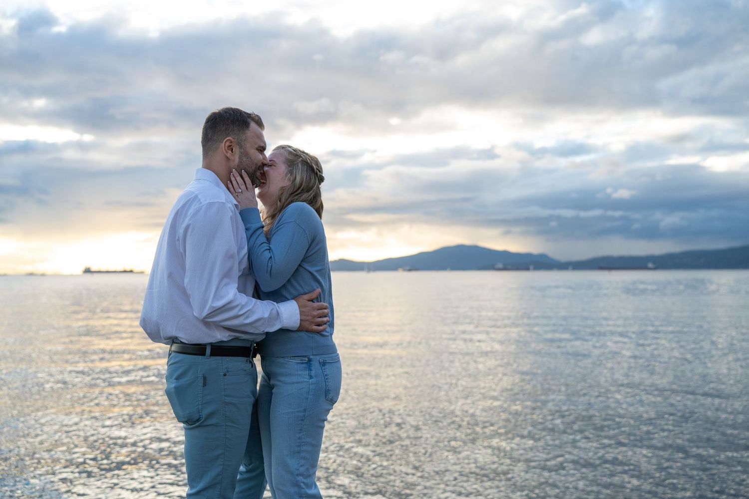Couple sharing a romantic kiss by the ocean at sunset with mountains and cloudy sky in the background.