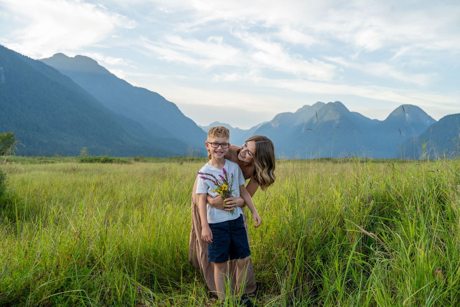 A mother kisses her young son in a lush green meadow with stunning mountain backdrop under a cloudy sky.