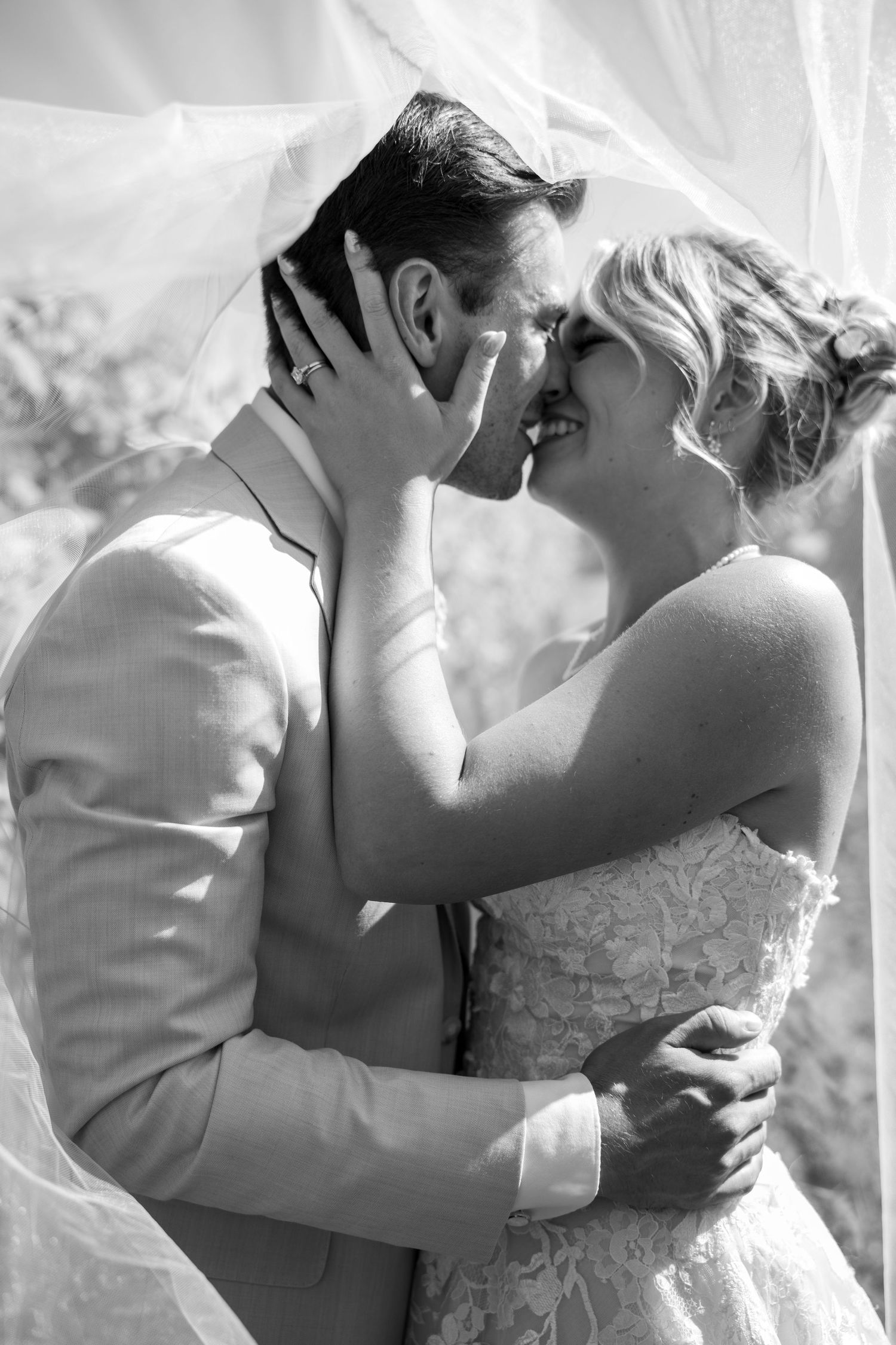 Black and white photo of a bride and groom sharing a romantic kiss under a flowing veil on their wedding day.