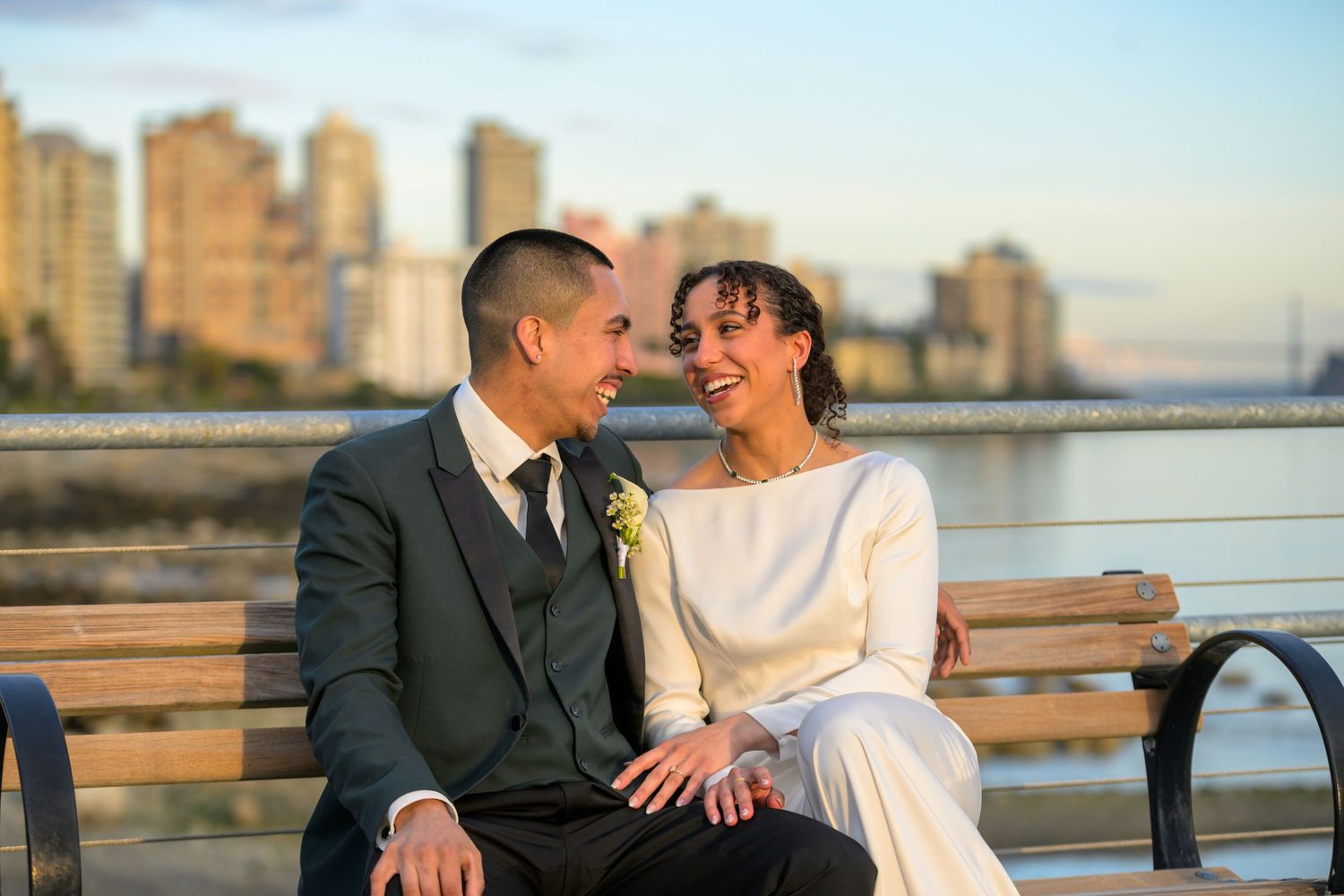 A romantic wedding couple shares a joyful moment on a bench with city skyline in the background at sunset.