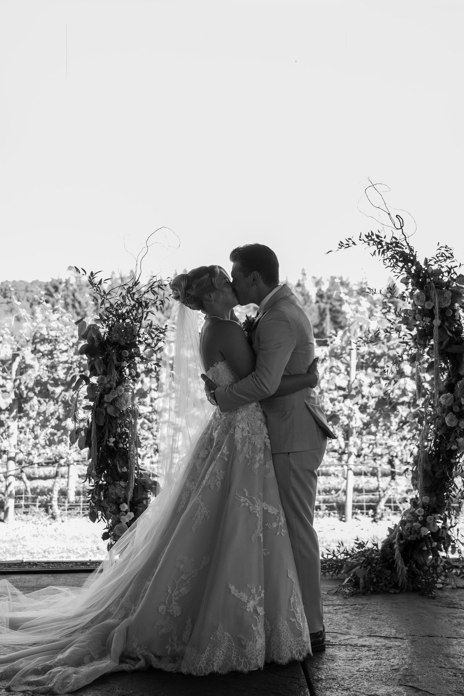 Black and white photo of a bride and groom sharing their first kiss at an outdoor vineyard wedding ceremony.