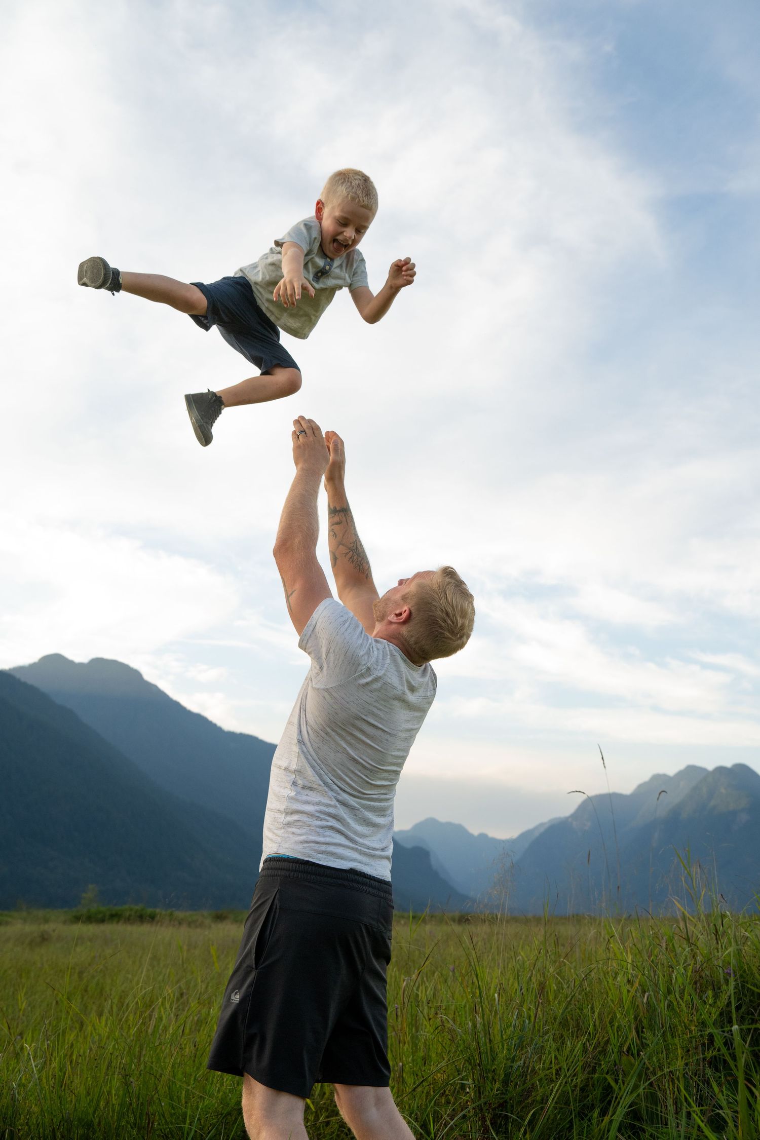 Father joyfully tosses young child into the air outdoors with scenic mountain range and cloudy sky in background.