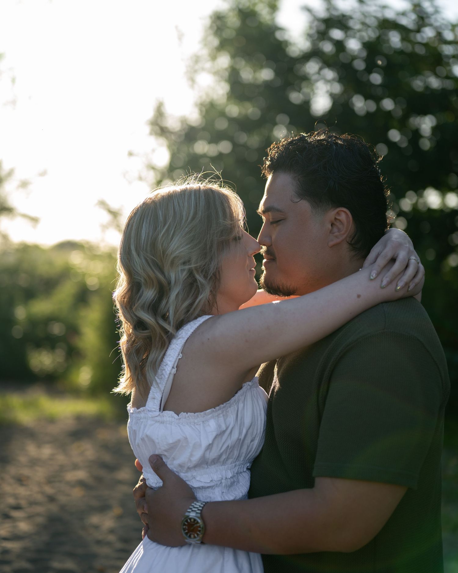A couple shares a romantic kiss outdoors at golden hour, surrounded by lush green trees in a warm, sunlit setting.