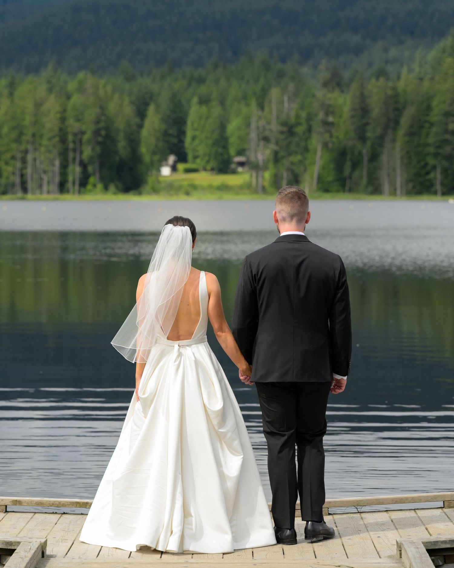 Bride and groom hold hands on a lakeside dock, gazing at lush green forest and mountain views on their wedding day.