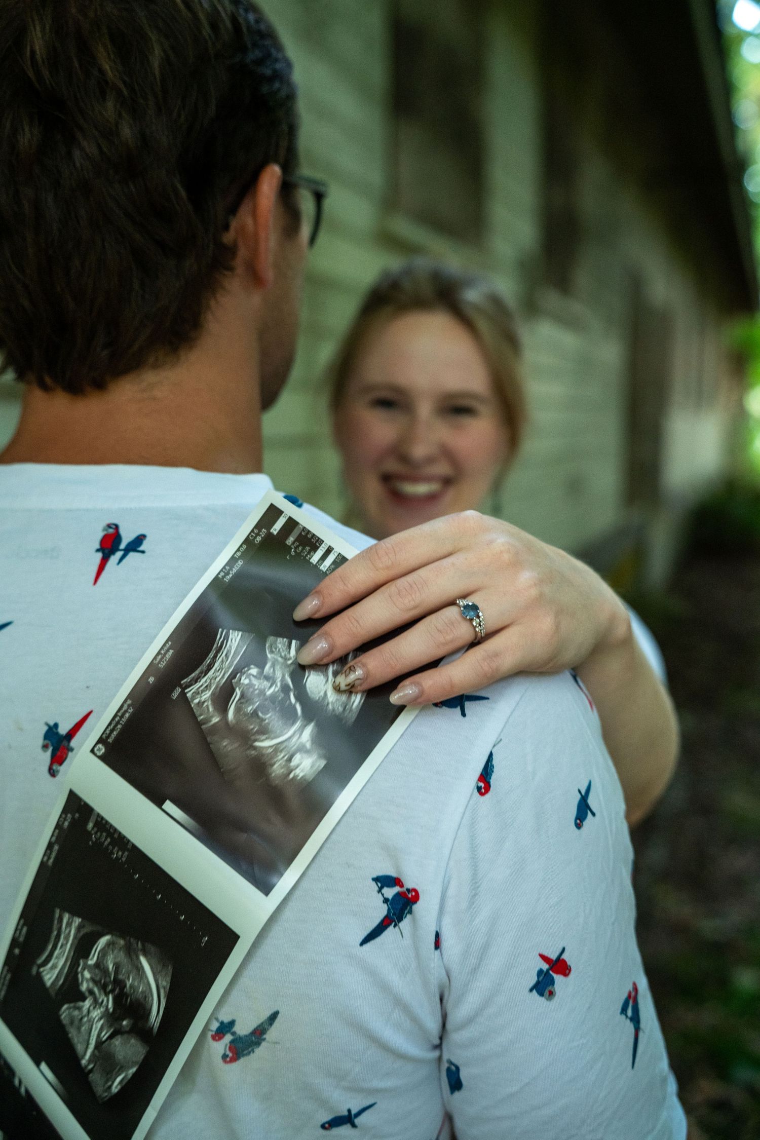 Smiling blonde woman holds ultrasound photos on man's shoulder, wearing engagement ring, announcing pregnancy outdoors.