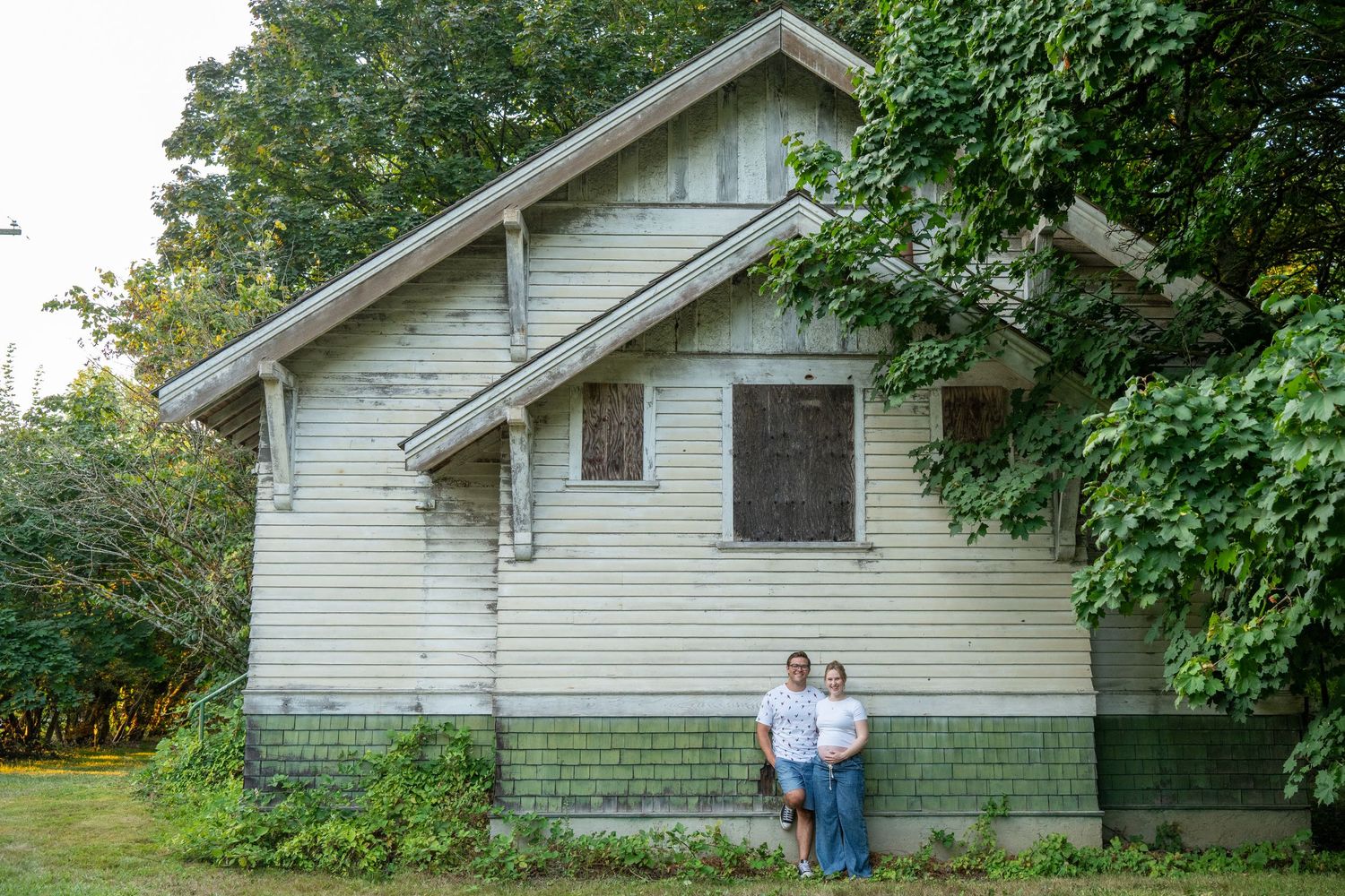 Two people stand in front of a weathered white clapboard house surrounded by lush green trees.