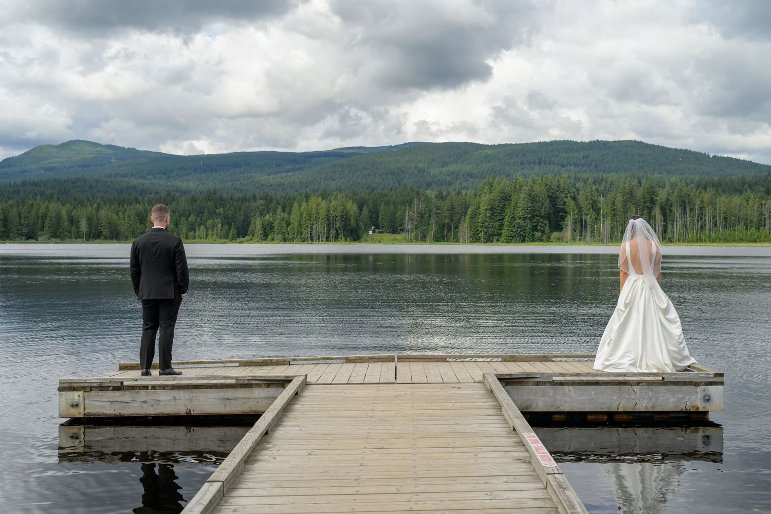 Bride and groom stand apart on a wooden dock overlooking a serene mountain lake surrounded by evergreen forests.