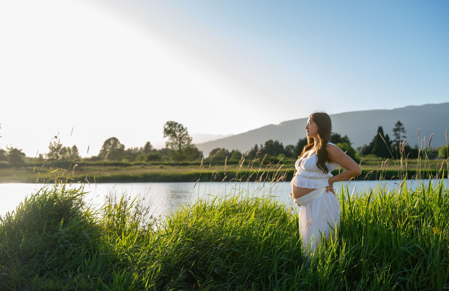 Pregnant woman in white dress standing by a serene lake with lush green grass and mountain backdrop in golden sunlight.