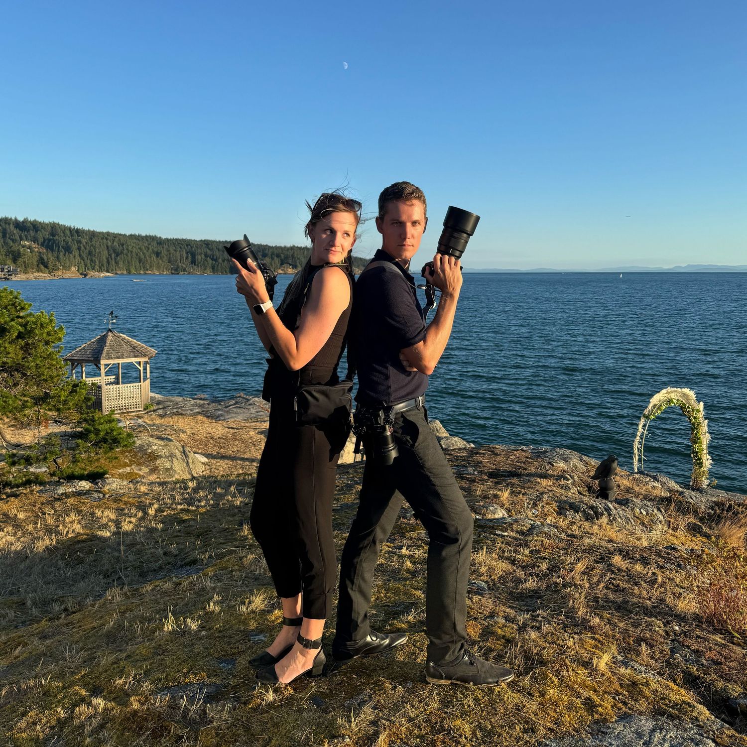 Two photographers holding cameras on coastal cliff with gazebo and ocean view. Sunset view from Bowen Island