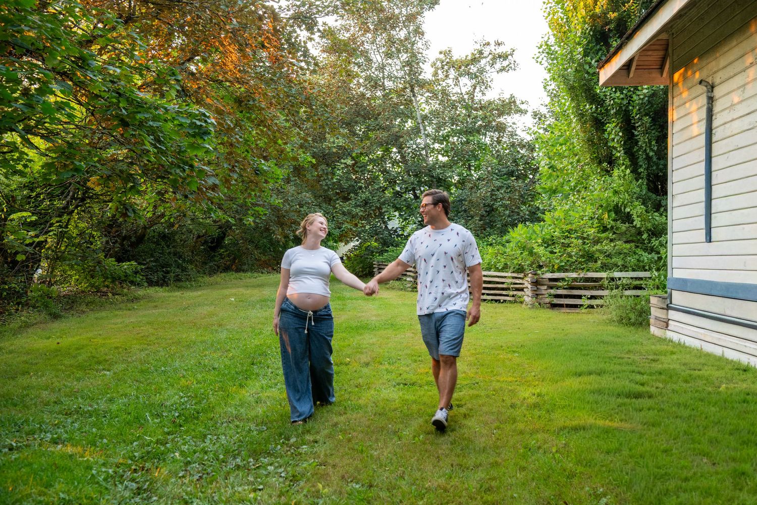 Pregnant couple holding hands walking on green lawn beside house surrounded by lush trees.