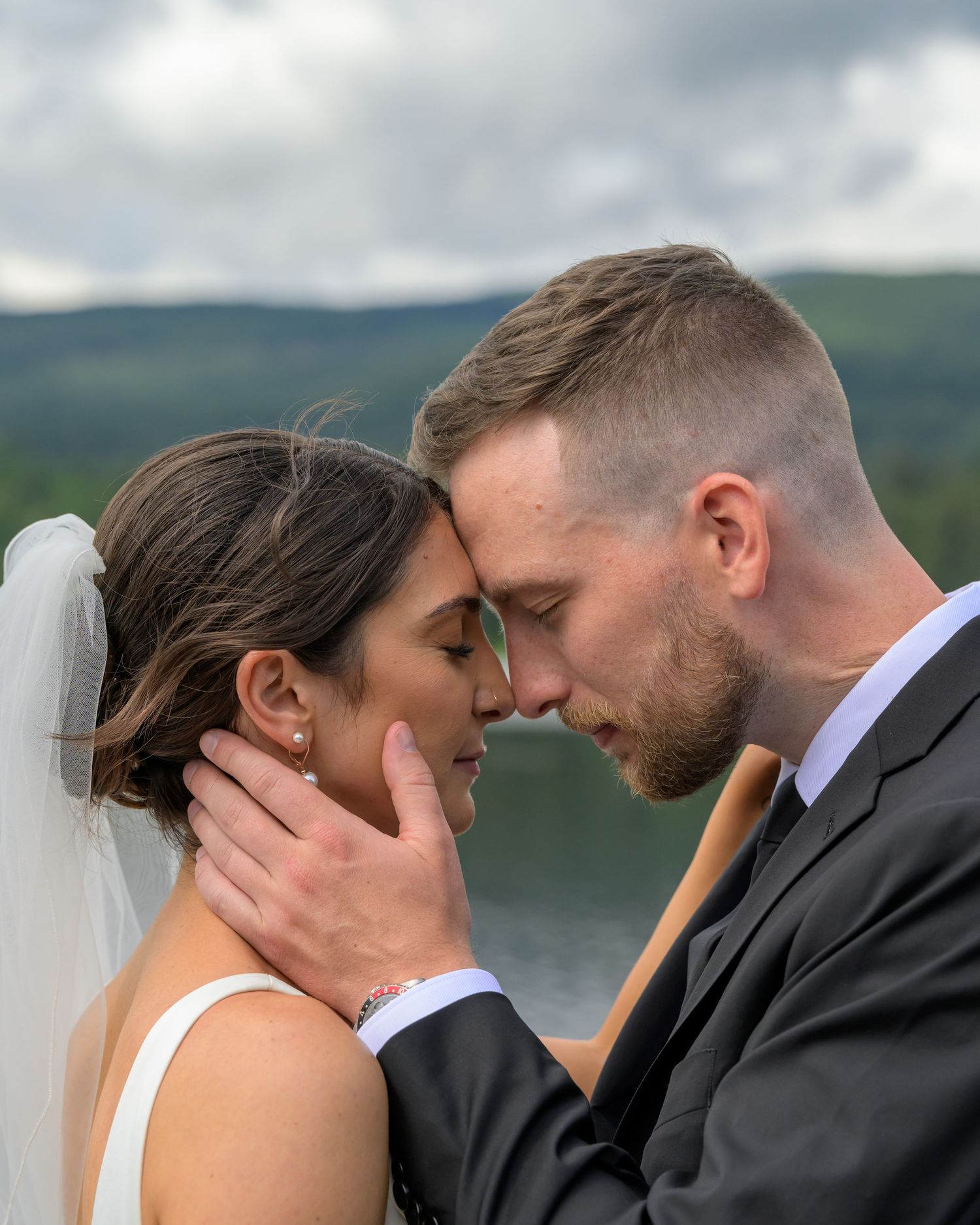 Romantic close-up of bride and groom touching foreheads tenderly with a scenic lake backdrop.