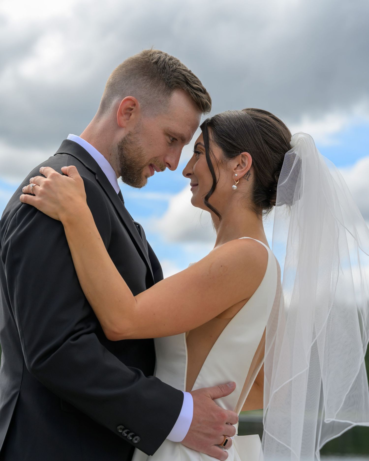 A bride and groom share an intimate moment, foreheads touching, dressed in wedding attire under a cloudy sky.