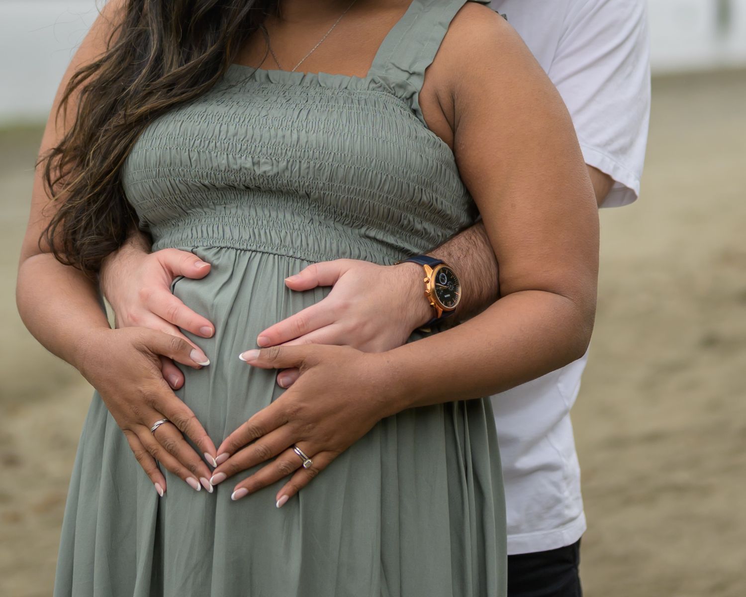 Pregnant woman in sage green dress cradling baby bump with partner embracing her from behind outdoors.
