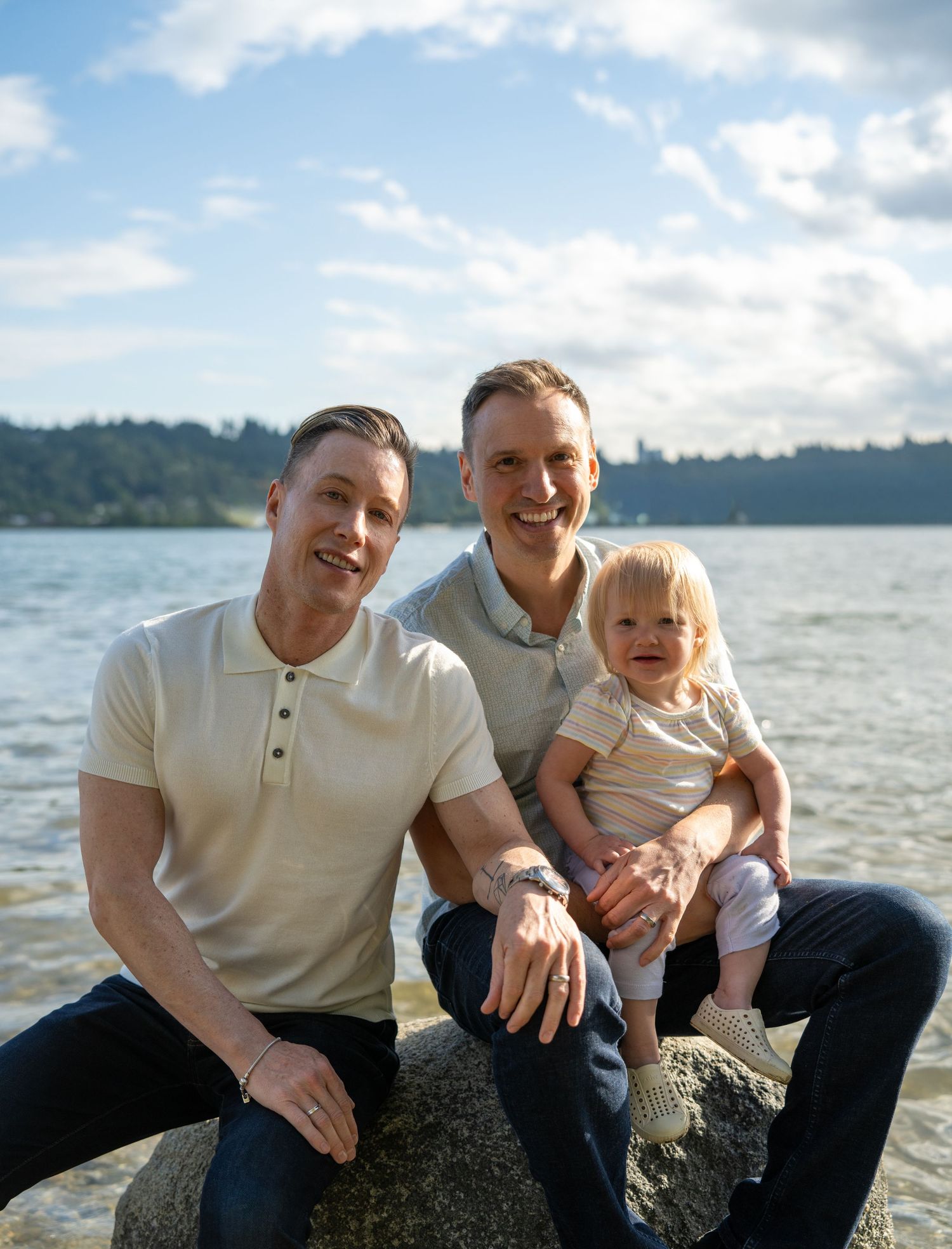 Two men and a toddler smile together on a rocky shoreline with a lake and blue sky in the background.