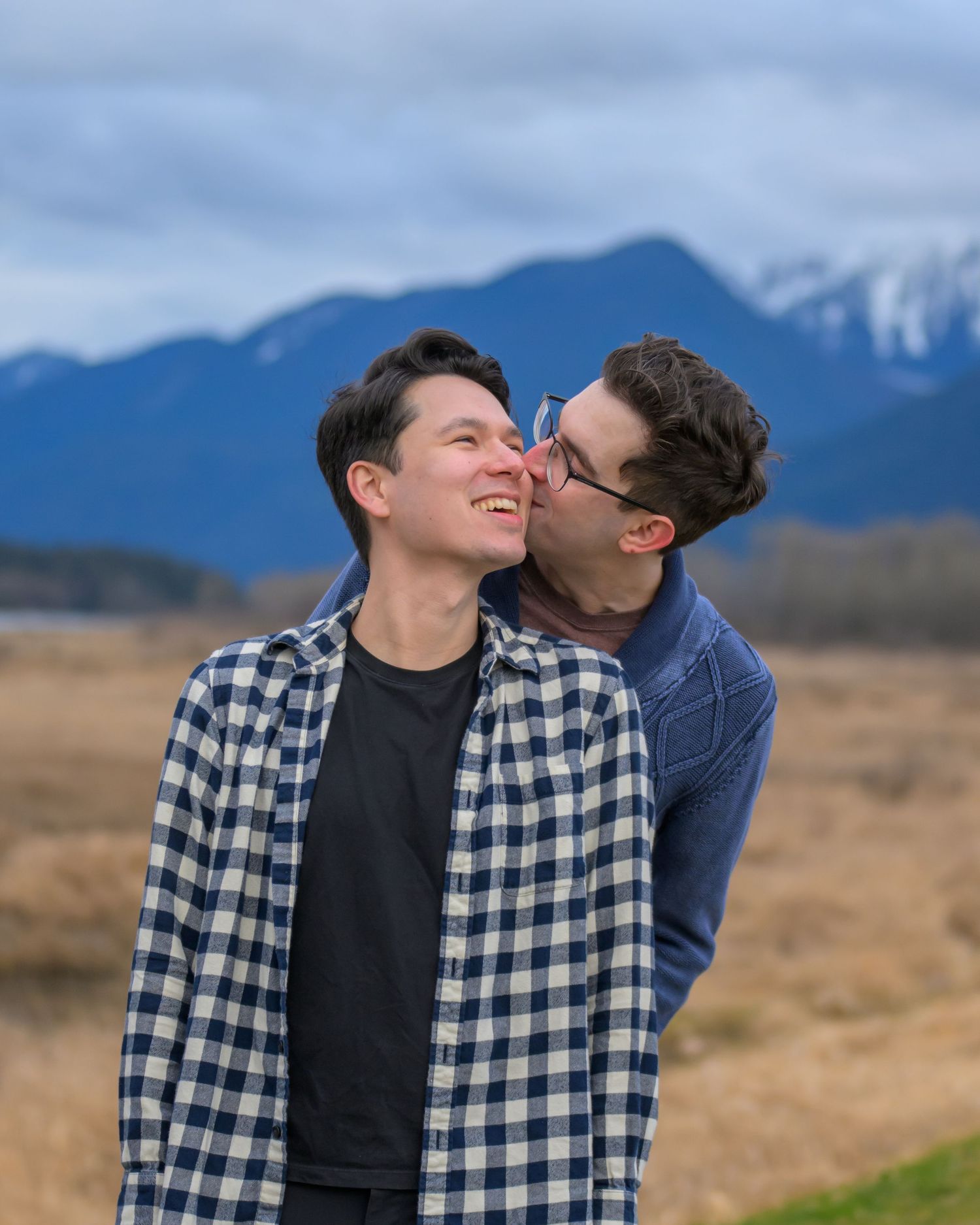 Couple sharing intimate moment in field with snow-capped mountains behind.