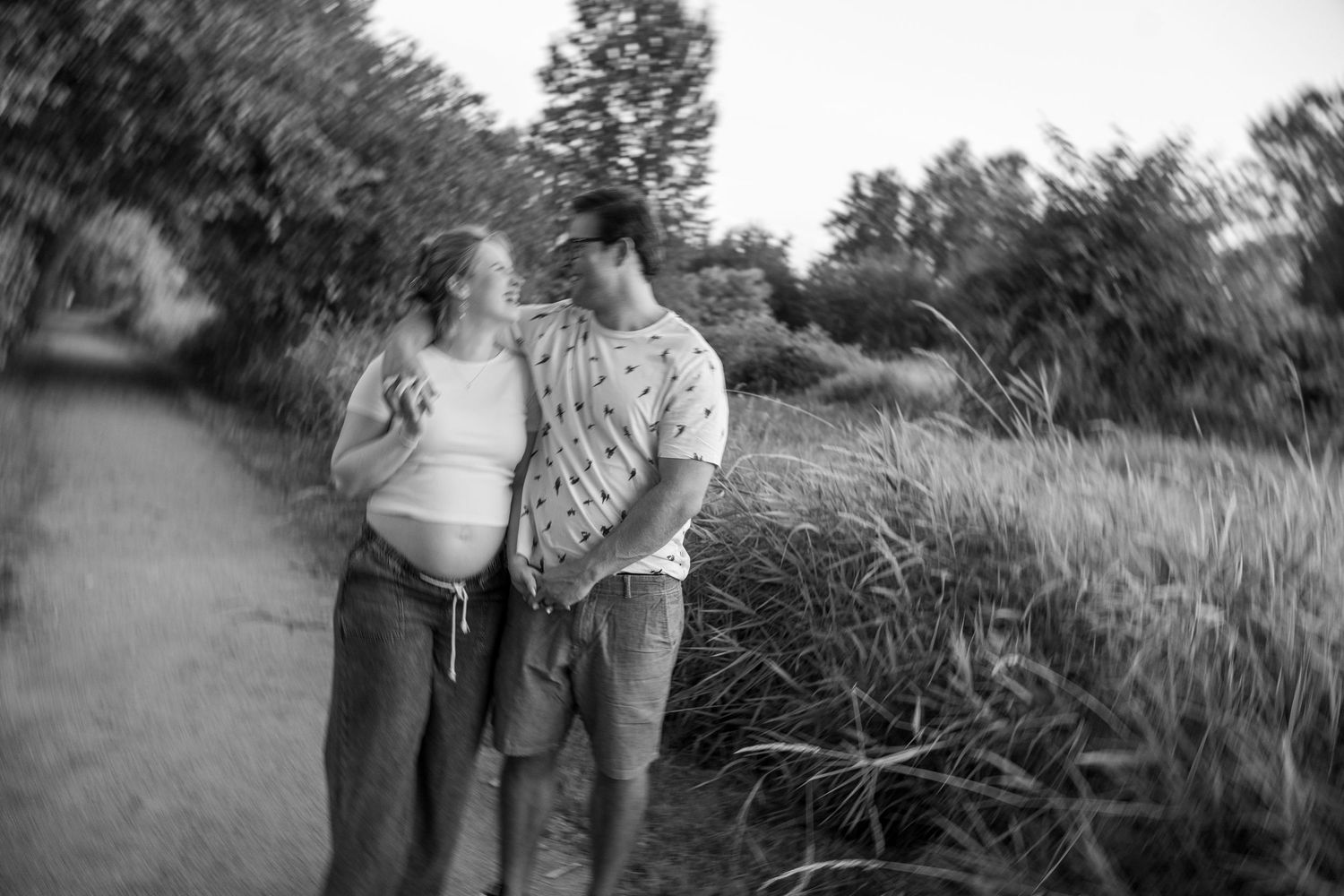 Black and white photo of a pregnant couple walking on a path surrounded by lush greenery.