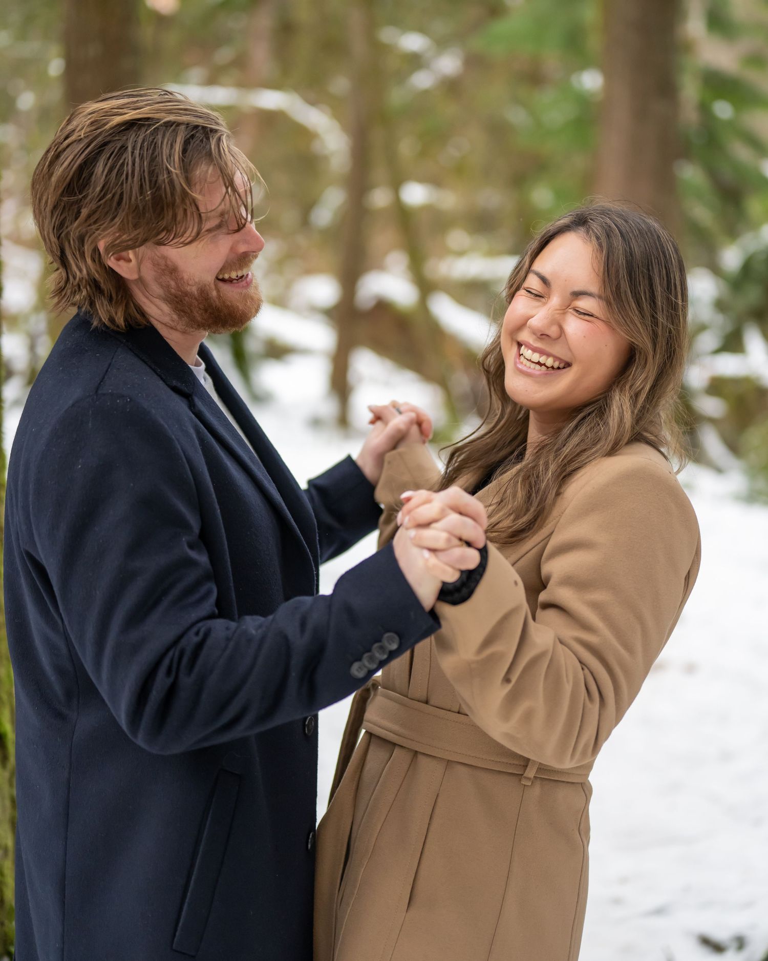 Couple in winter coats sharing joyful moments in snowy forest setting.