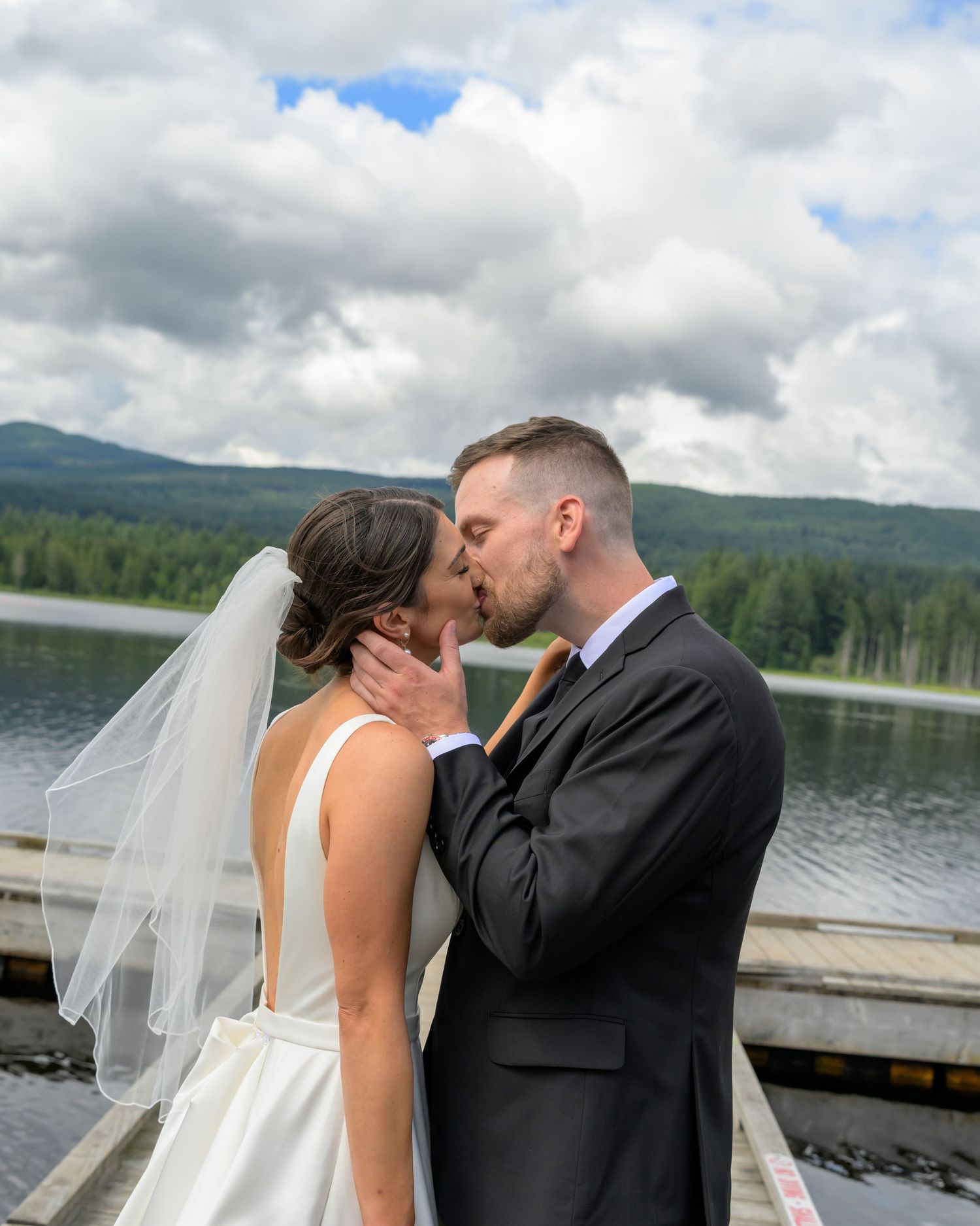 Newlyweds share a romantic kiss on a dock with a scenic lake and mountains behind.