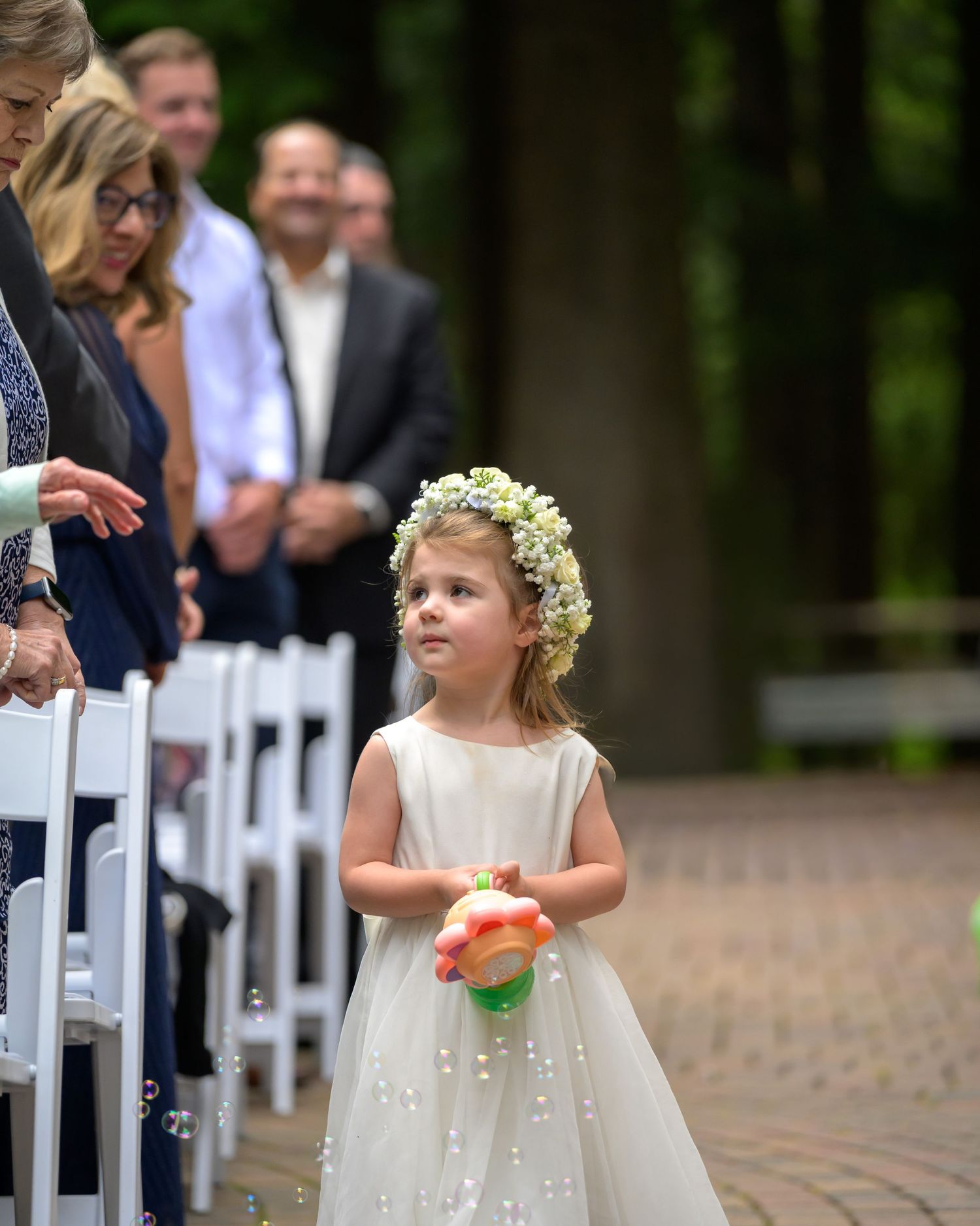 Flower girl in white dress and floral crown blowing bubbles down wedding aisle at outdoor ceremony.