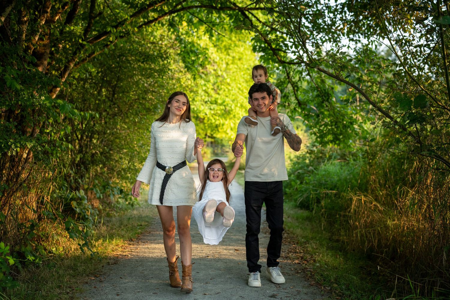 Family walking together holding hands on a tree-lined path during a sunny summer afternoon. Colony Farms park in Port Coquitlam