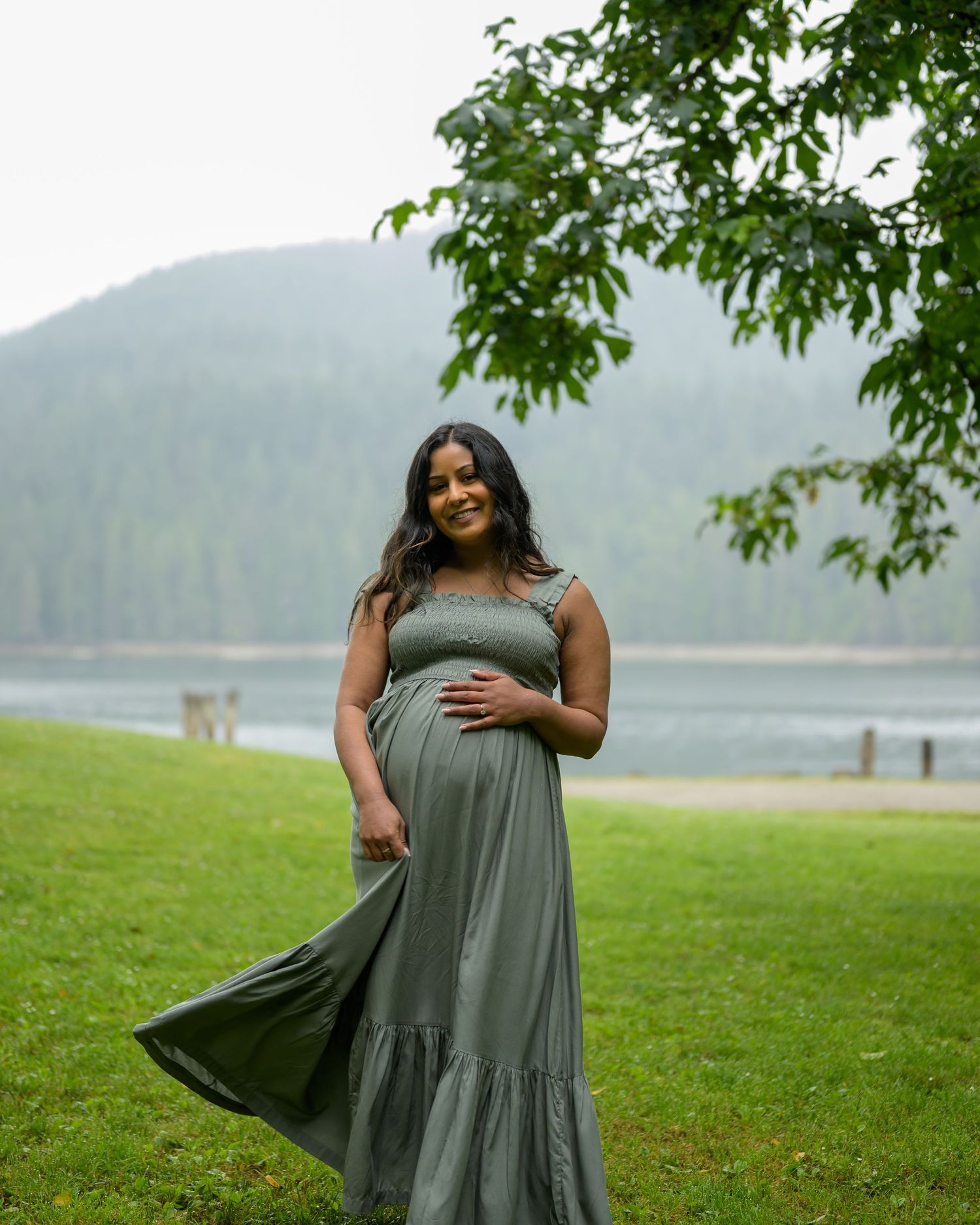 Smiling pregnant woman in olive green maxi dress standing on grass near a lake with mountain backdrop.