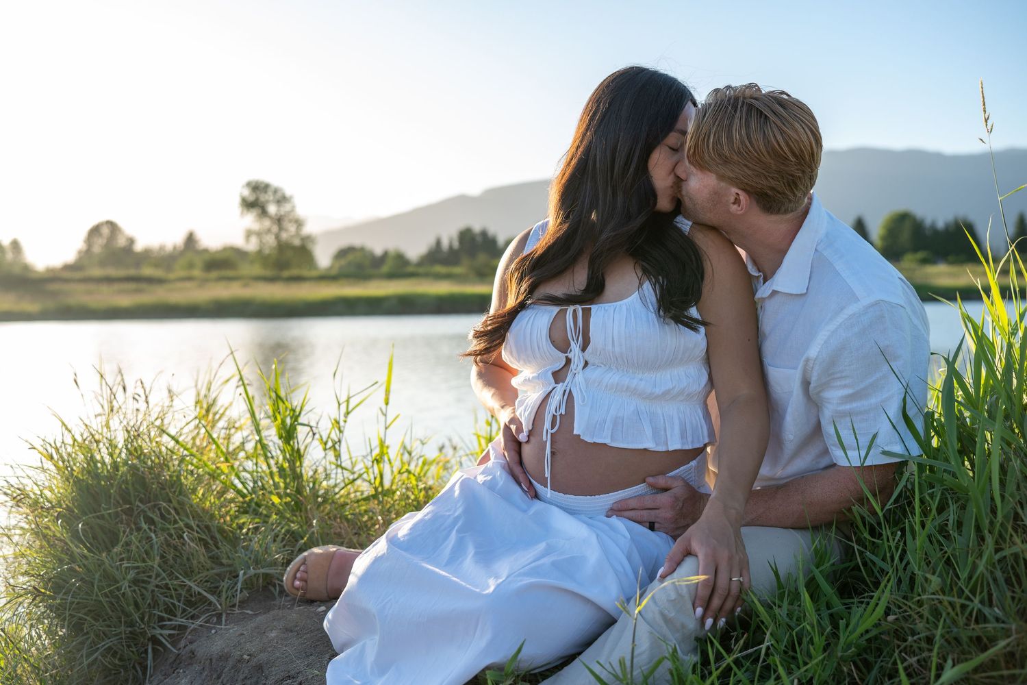 Couple sharing a romantic moment by a serene lake at sunset, woman in white dress with baby bump visible.