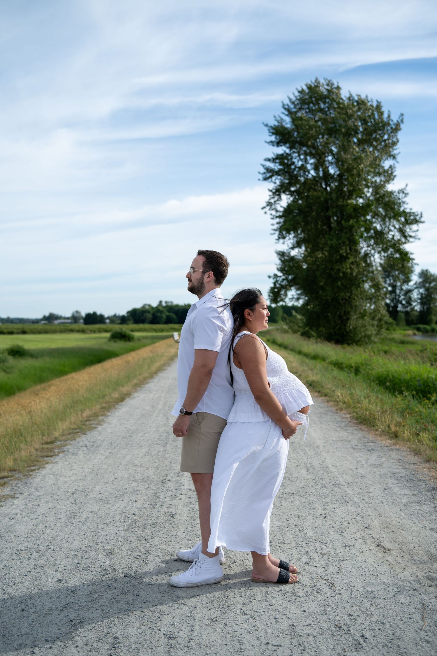Pregnant couple standing back-to-back on a rural gravel road surrounded by green fields and trees under a blue sky.
