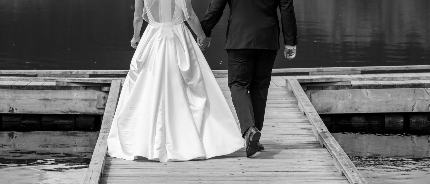 Bride and groom walking hand in hand on a wooden dock by the water in a black and white wedding photo.