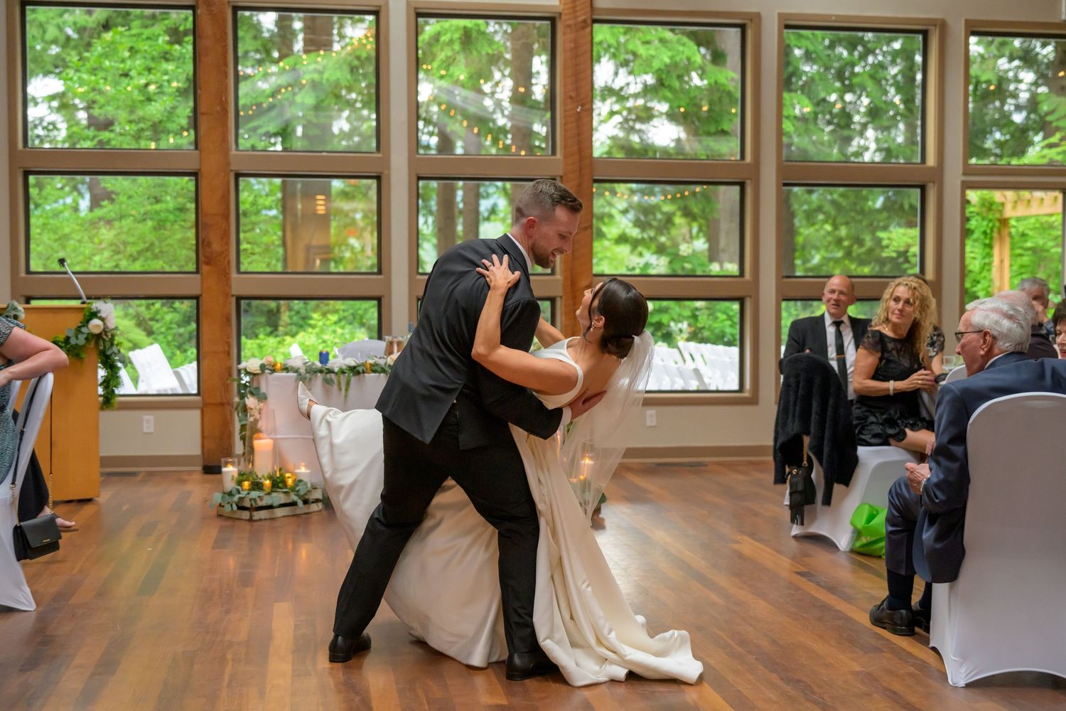 Bride and groom share romantic first dance dip in elegant wood-paneled wedding reception hall with guests watching.