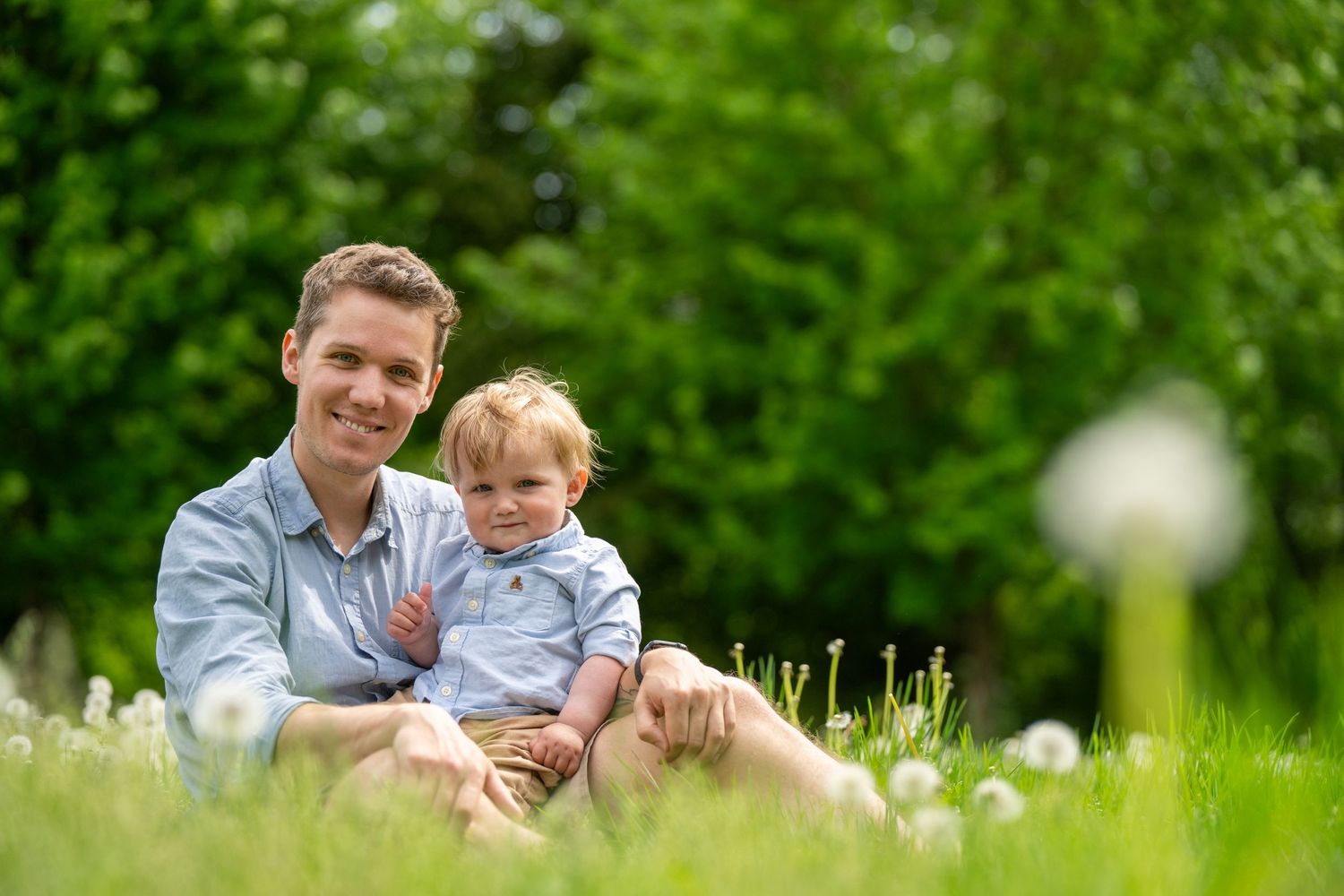A smiling father sits in a green meadow holding his toddler son, surrounded by dandelions on a sunny day.