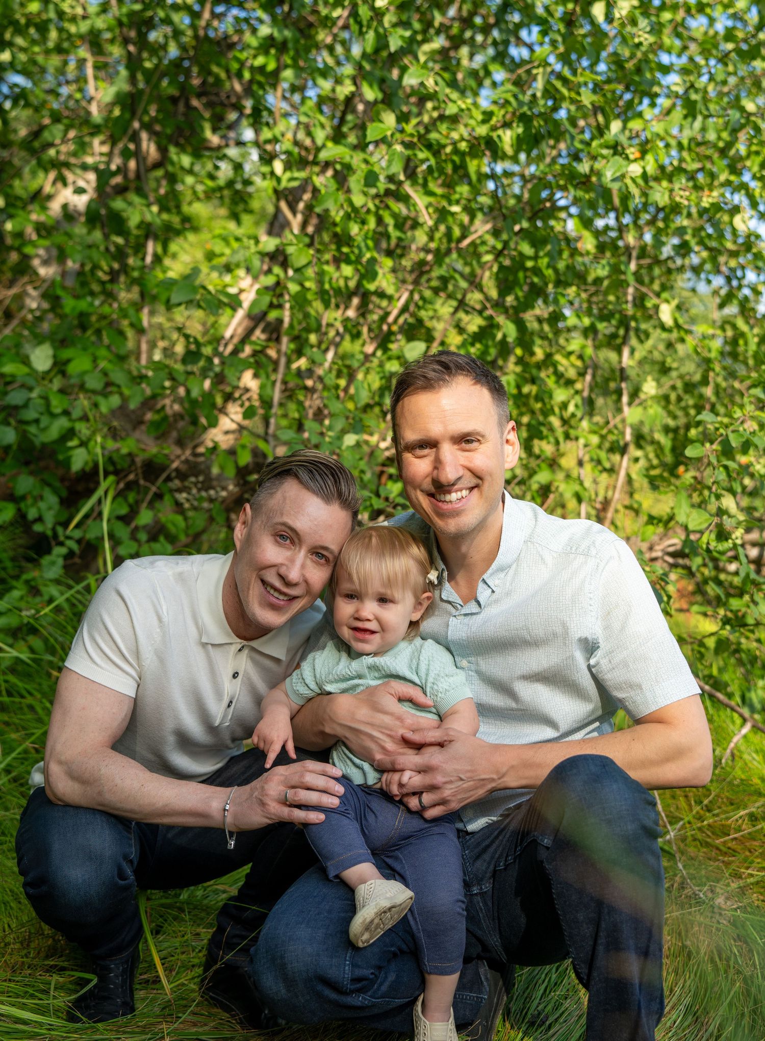 Two men and a toddler smile together outdoors in lush green nature setting for a family portrait photo.