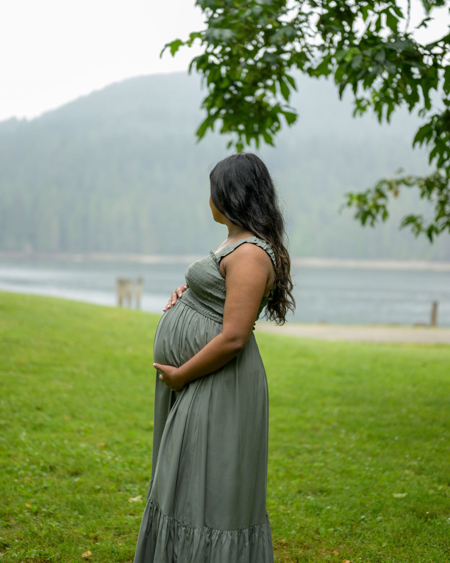 Pregnant woman in sage green maxi dress cradling her baby bump outdoors near a misty lake and green hills.