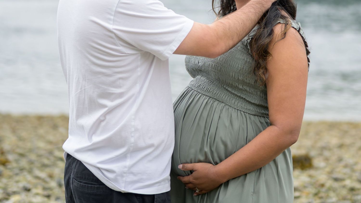 Couple sharing a tender moment on a rocky beach, man embracing pregnant woman in sage green maternity dress.