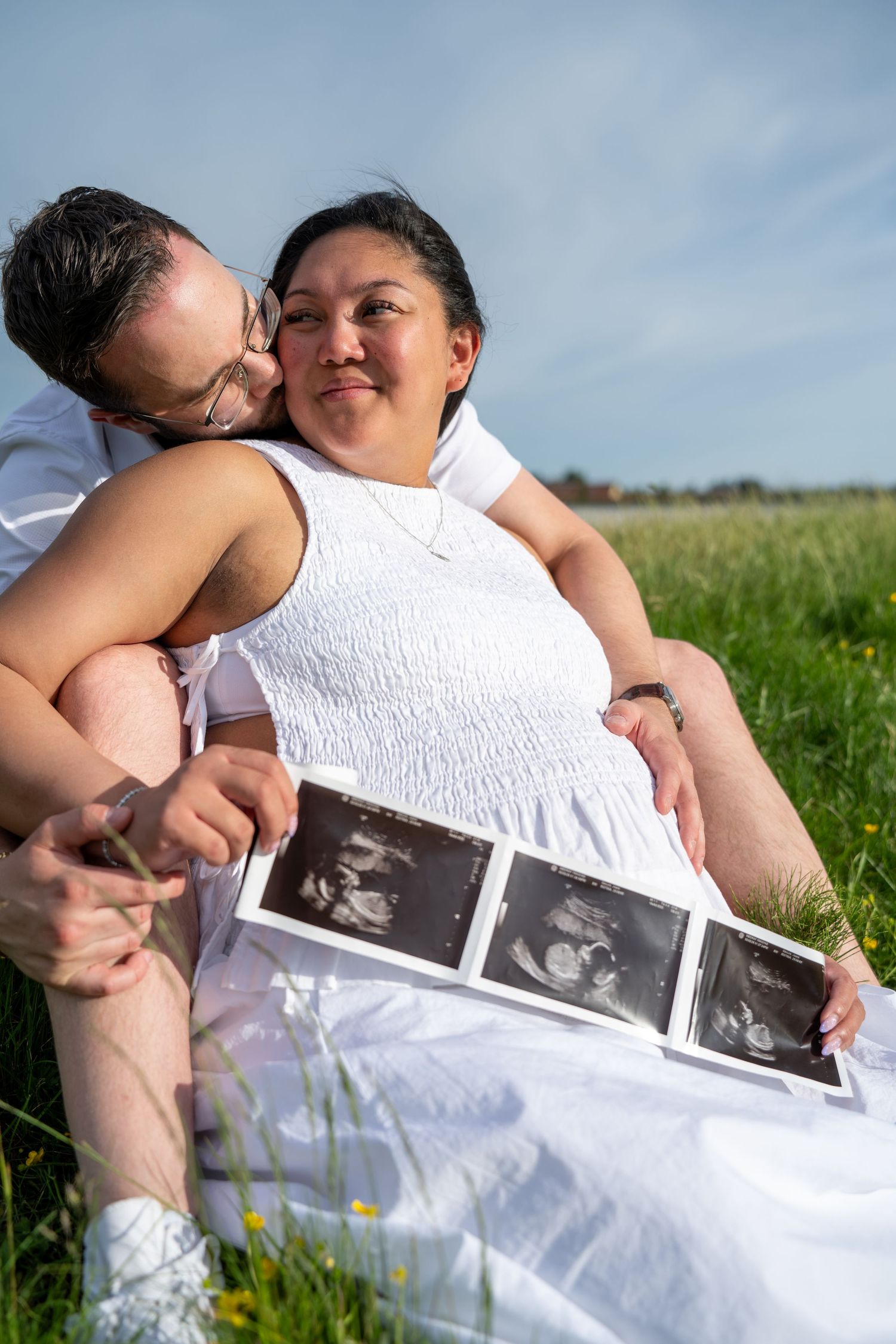 Couple sitting in green field, woman in white dress holding ultrasound photos while partner kisses her cheek on sunny day.