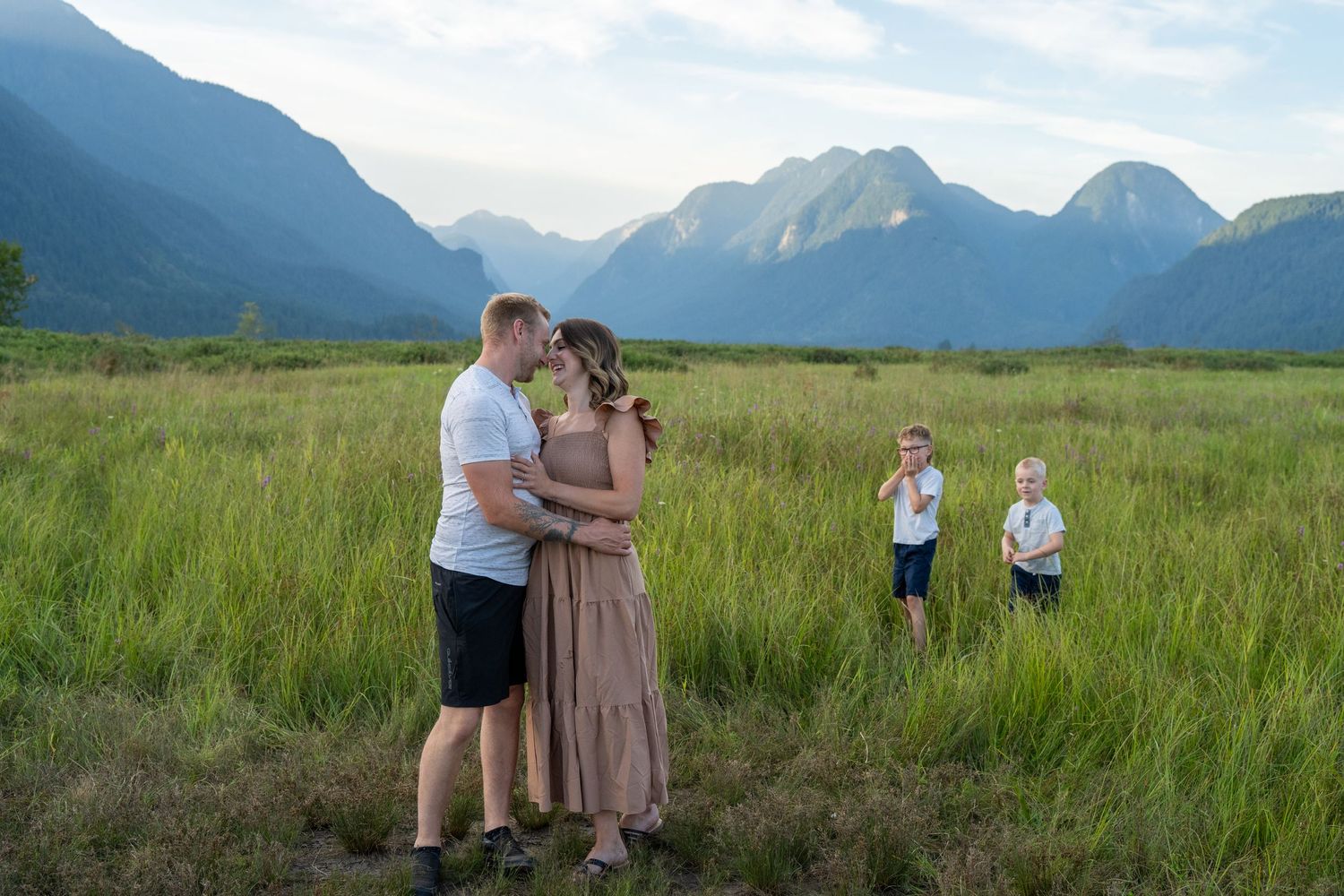 Family of four poses in lush green meadow with stunning mountain backdrop during golden hour outdoor photo session.