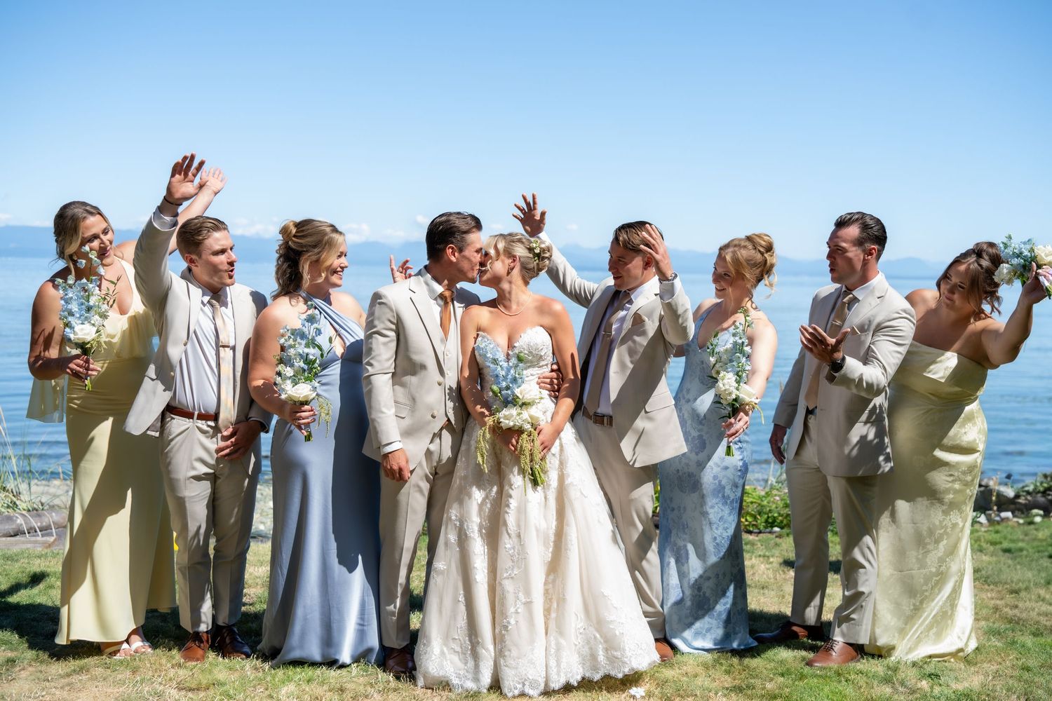 Wedding party celebrating outdoors by the ocean, bride and groom kissing while bridesmaids and groomsmen cheer around them.