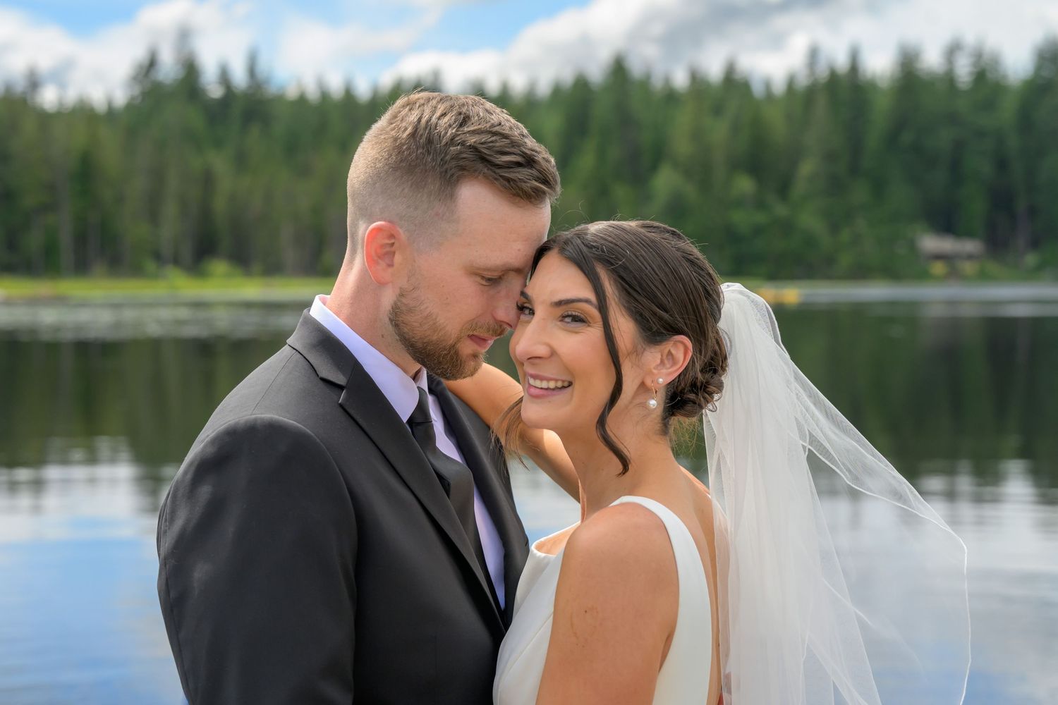 A bride and groom share a romantic moment by a serene lake surrounded by evergreen trees on their wedding day.