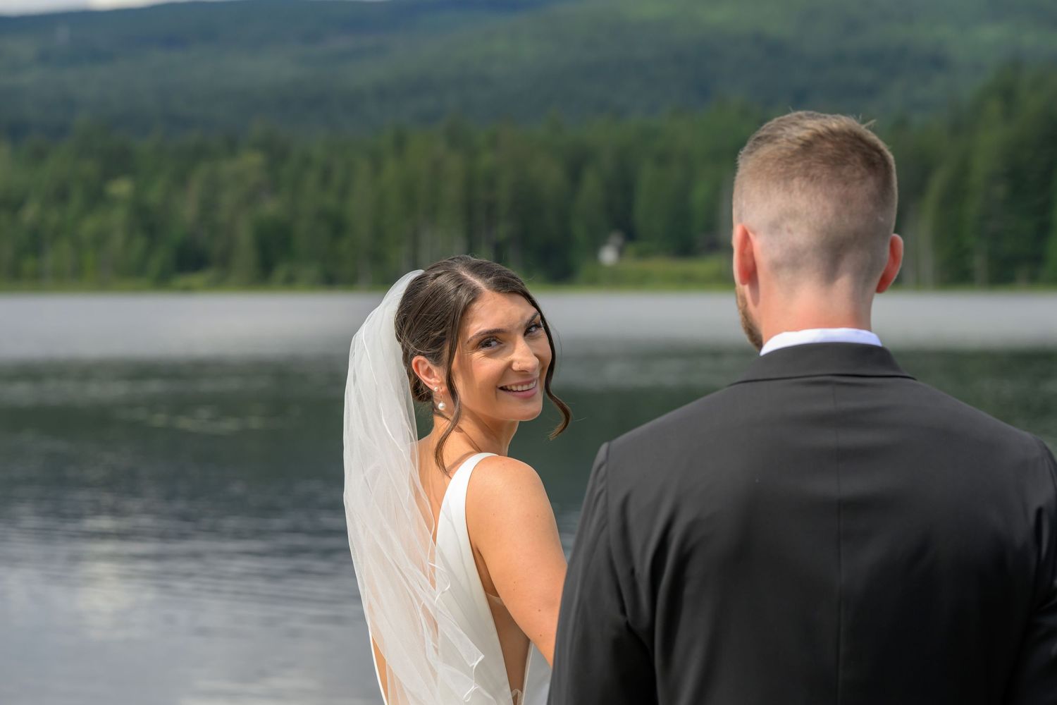 Bride in white gown and veil smiles at groom during outdoor lakeside wedding ceremony with forested mountains behind.