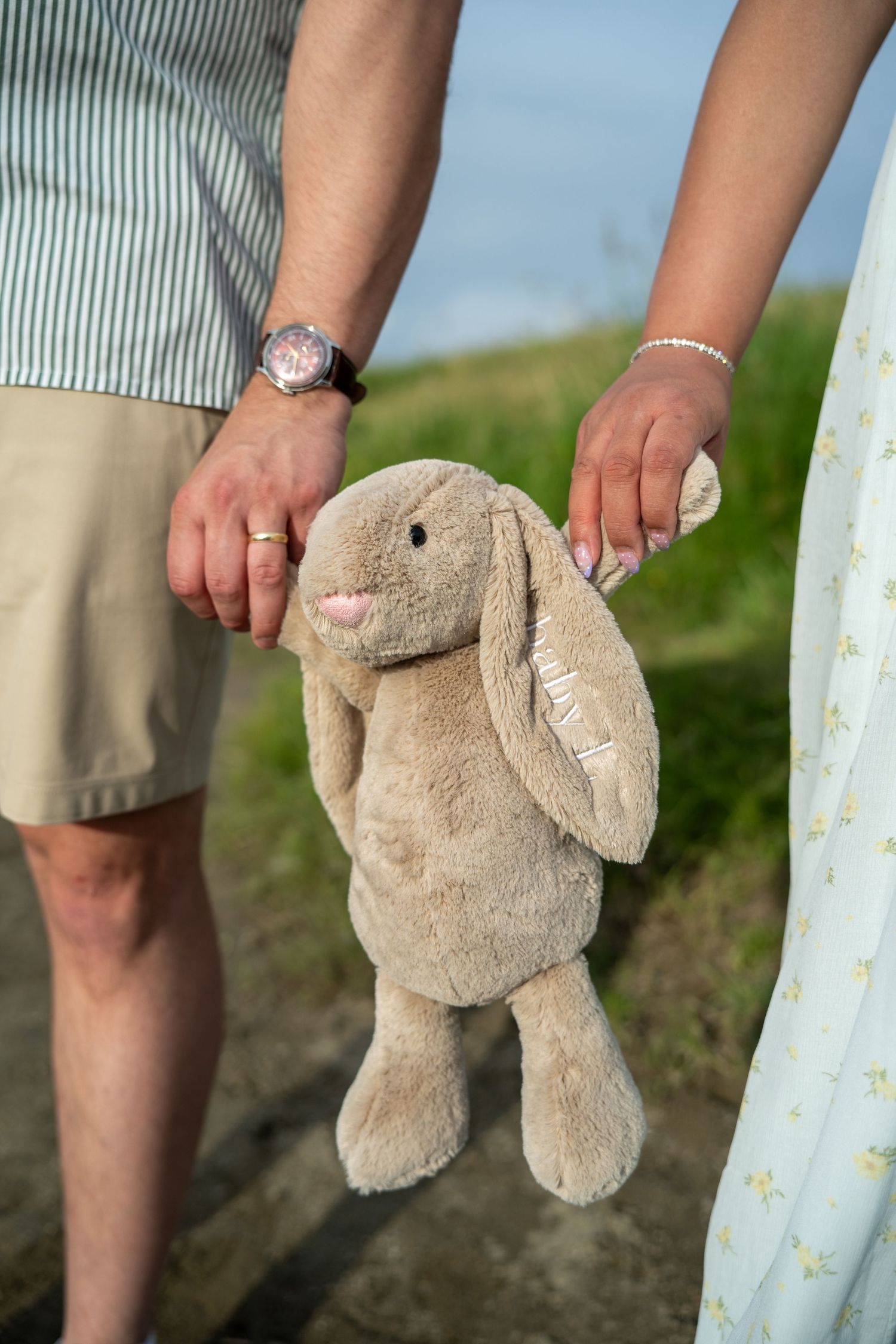 Couple holding a soft beige stuffed bunny toy together outdoors, symbolizing an upcoming baby announcement.