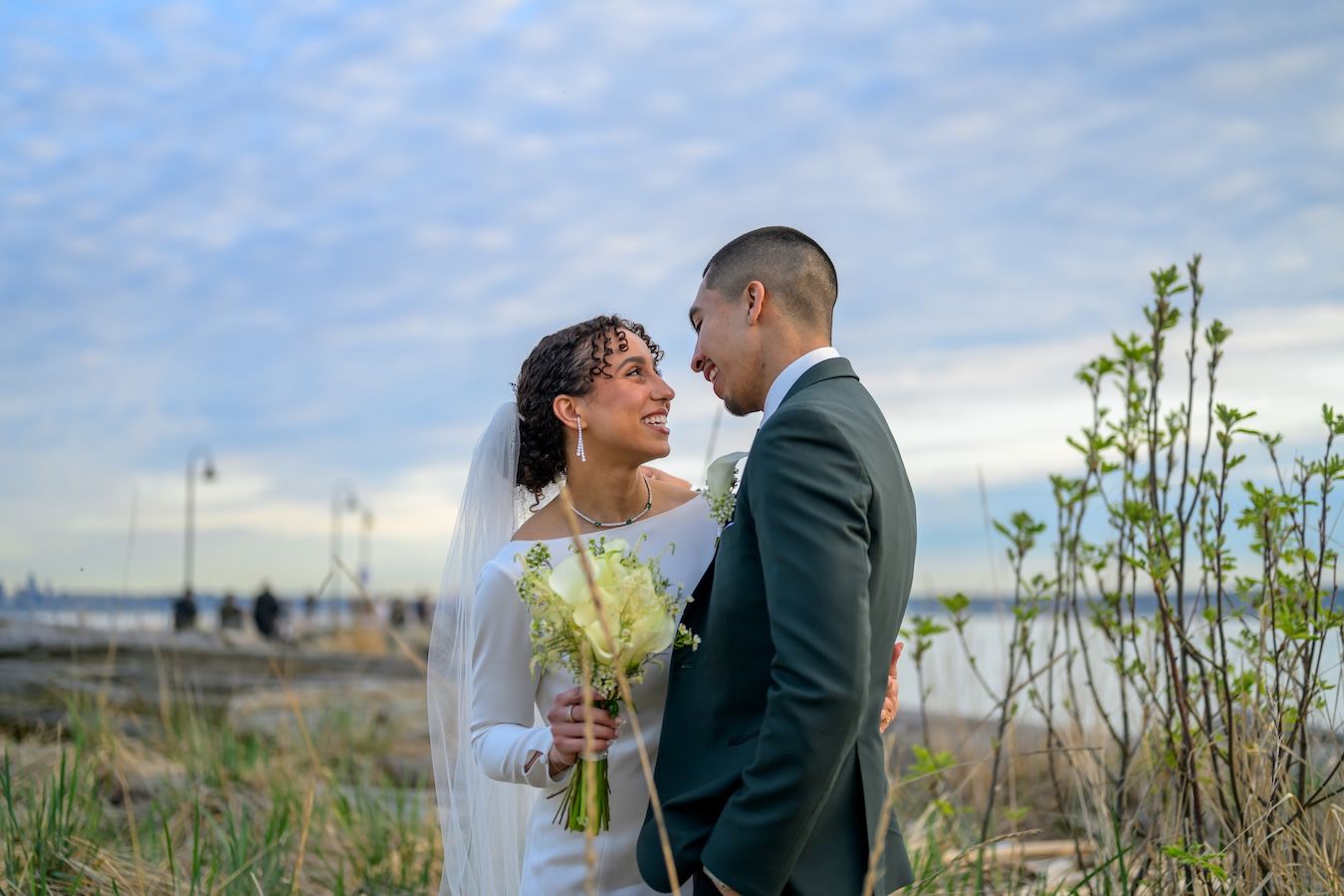 Couple in wedding attire embracing on a scenic rocky beach with mountains and clouds in the background.