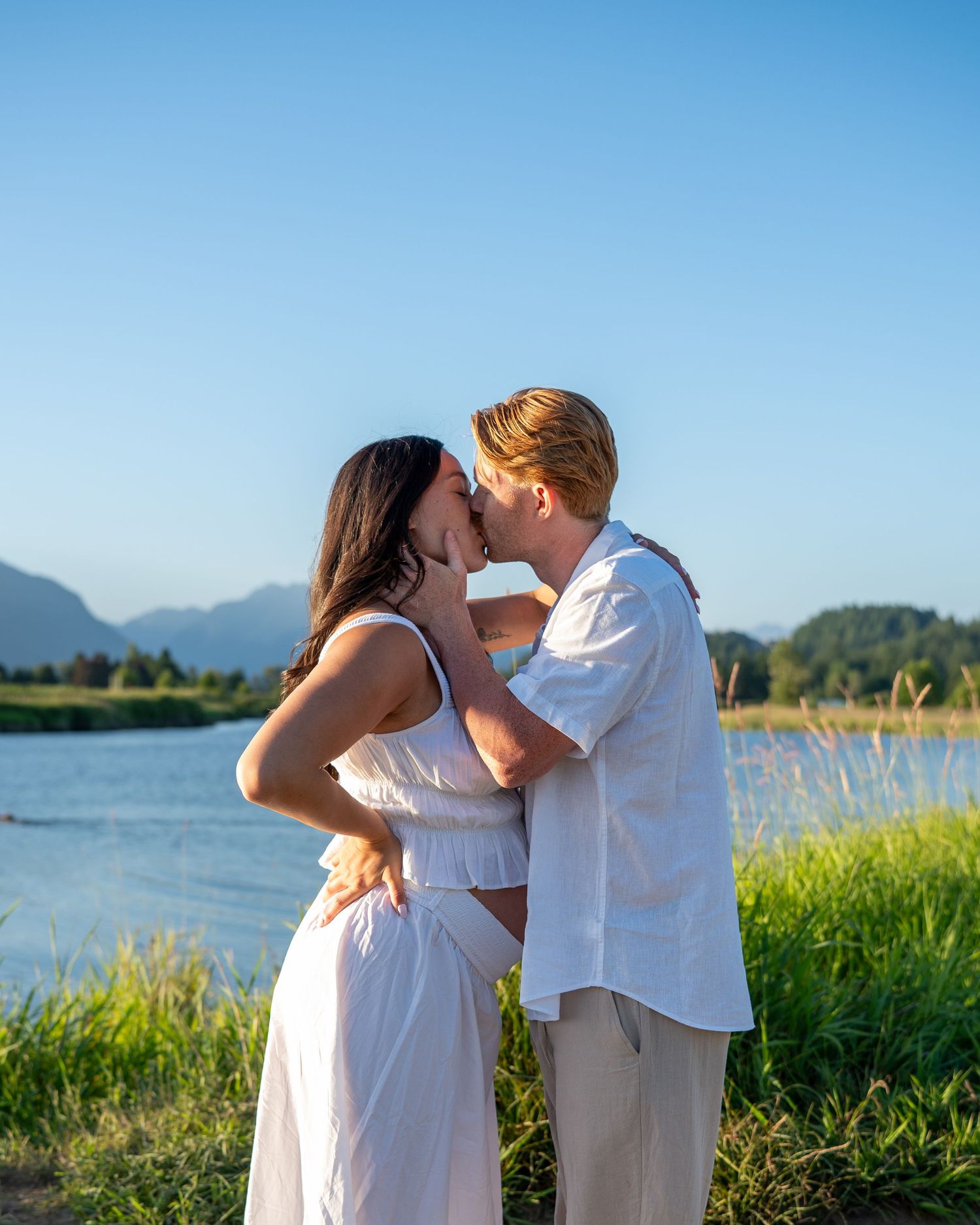 Couple kissing by a scenic lake with mountains and green grass under a clear blue sky.
