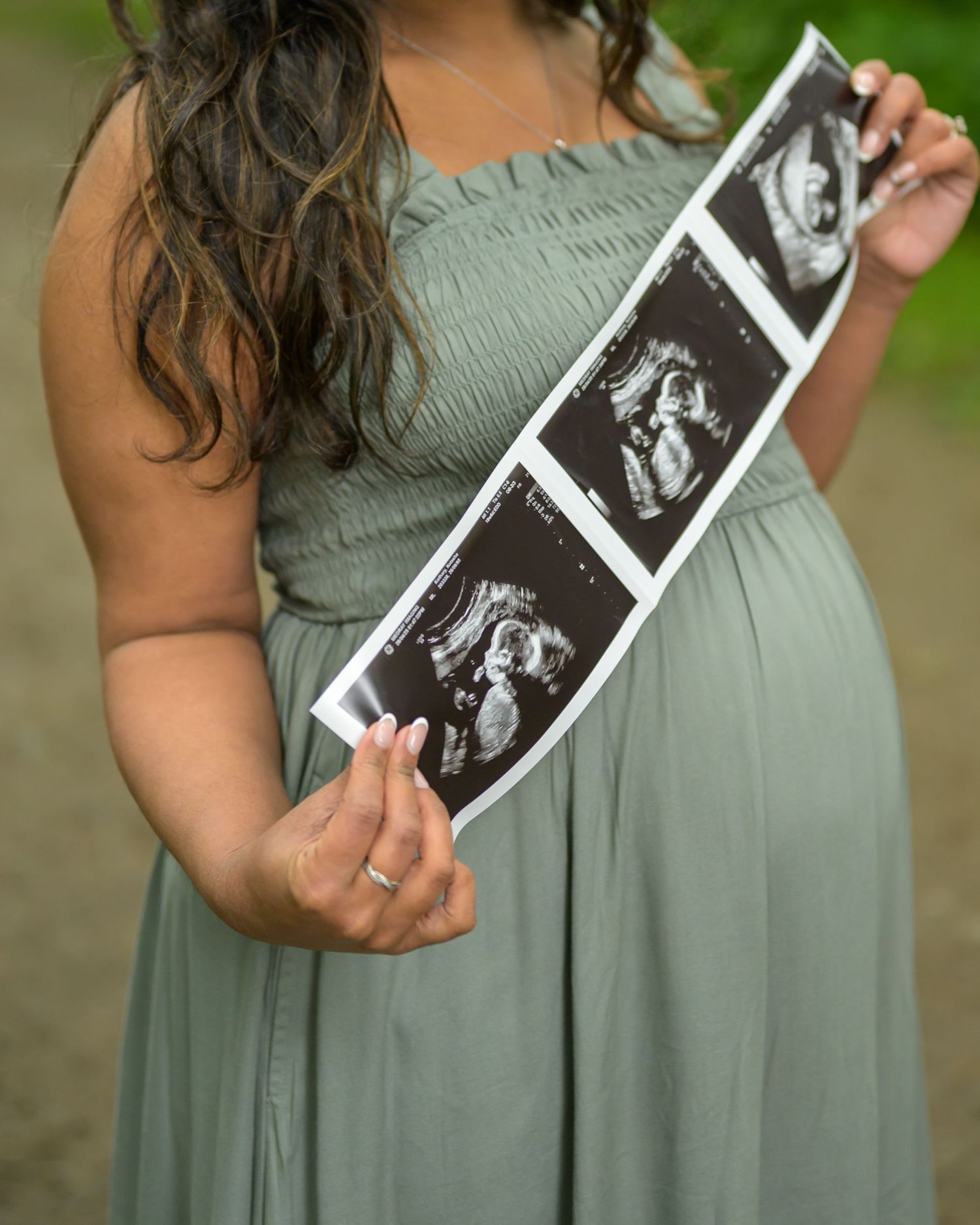 Pregnant woman in sage green dress holding ultrasound photos outdoors during maternity photo session.