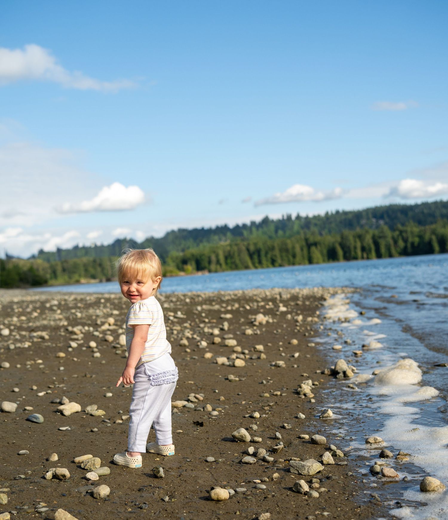 A toddler with blonde hair stands on a rocky beach shore, smiling, with blue water and green trees in the background.