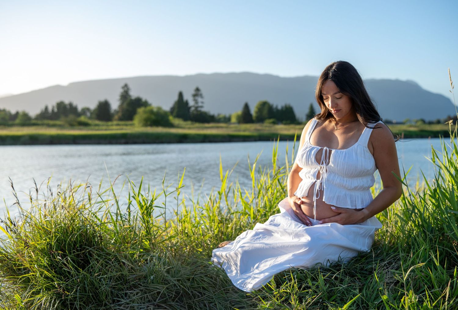 Pregnant woman in white dress sitting by a serene lake surrounded by lush green grass and mountains in sunlight.